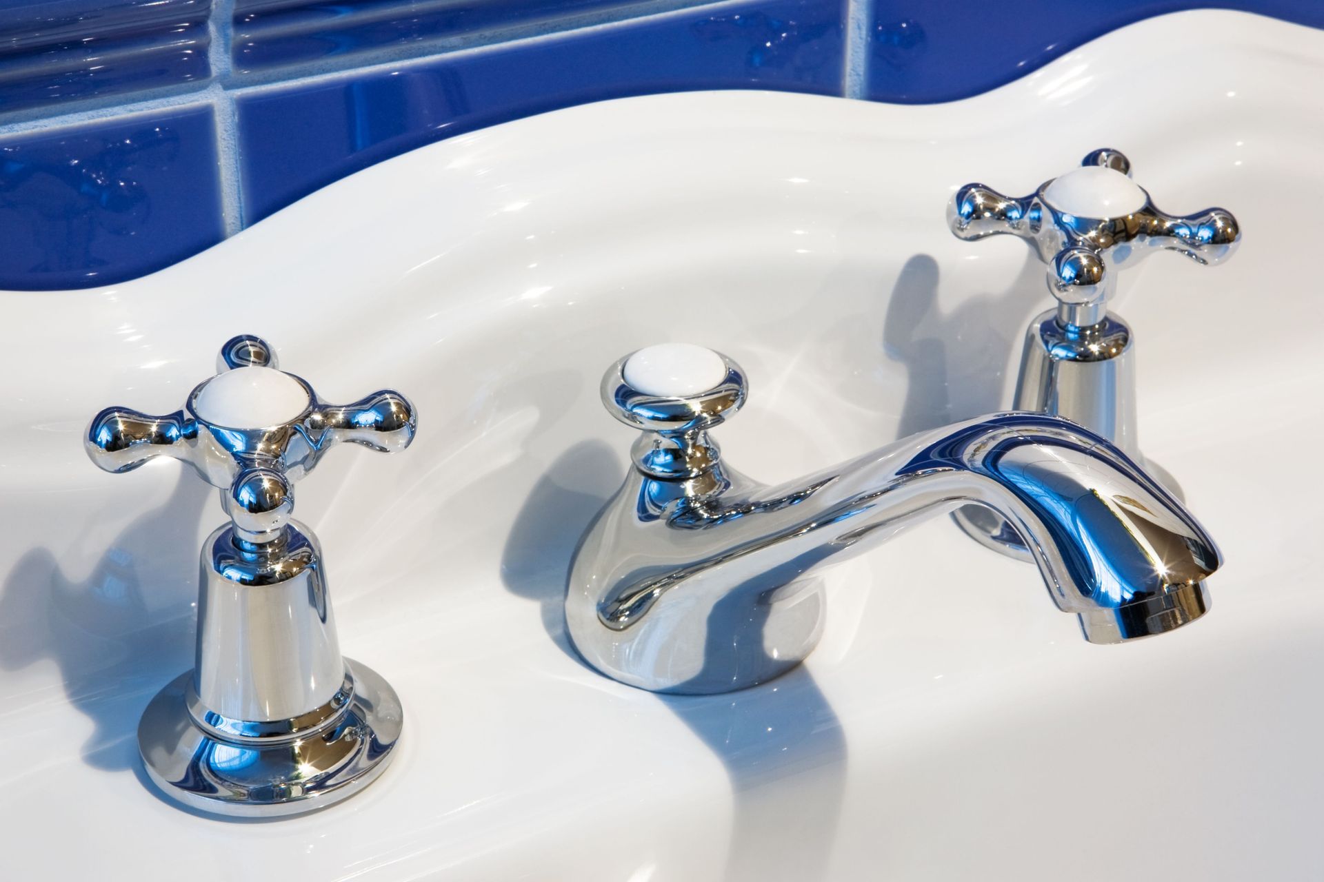 A chrome three-piece faucet set with white accents mounted on a white porcelain sink against a blue tiled wall.