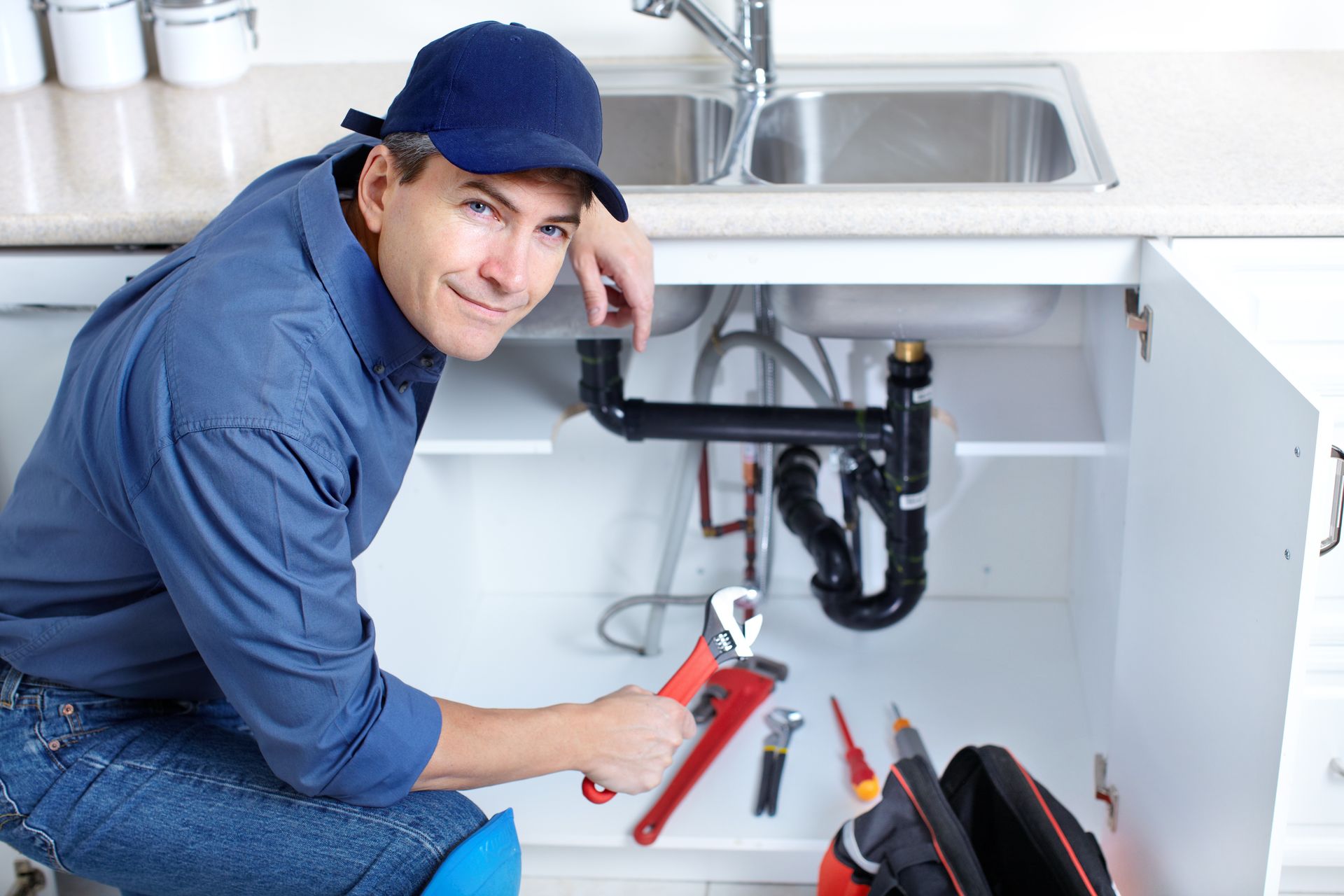 A smiling plumber in a blue uniform kneels under a kitchen sink, holding a wrench with tools nearby.
