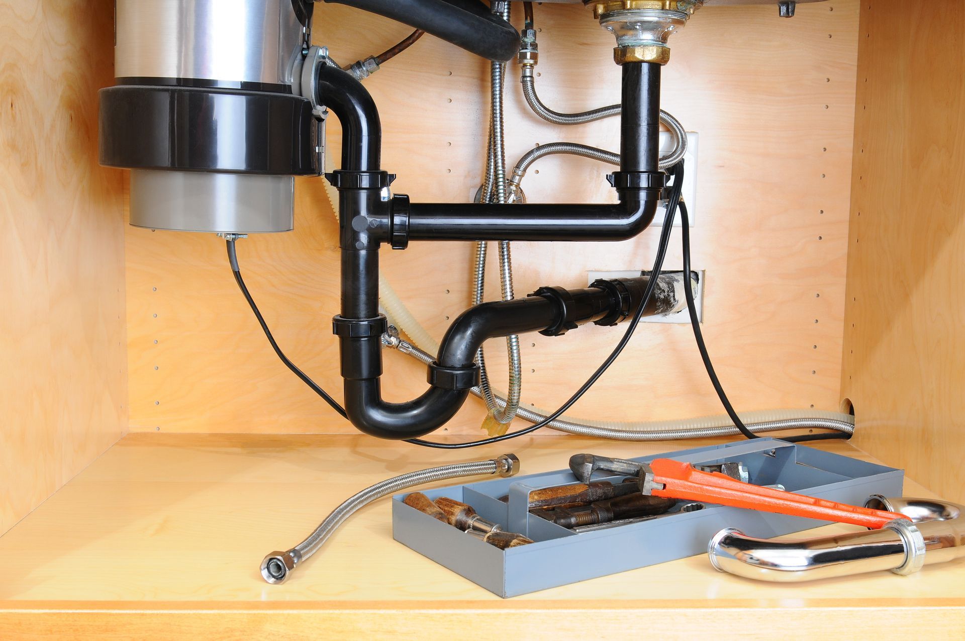 The underside of a kitchen sink, showing black plastic plumbing, a garbage disposal, tools, and metal pipes in a cabinet.