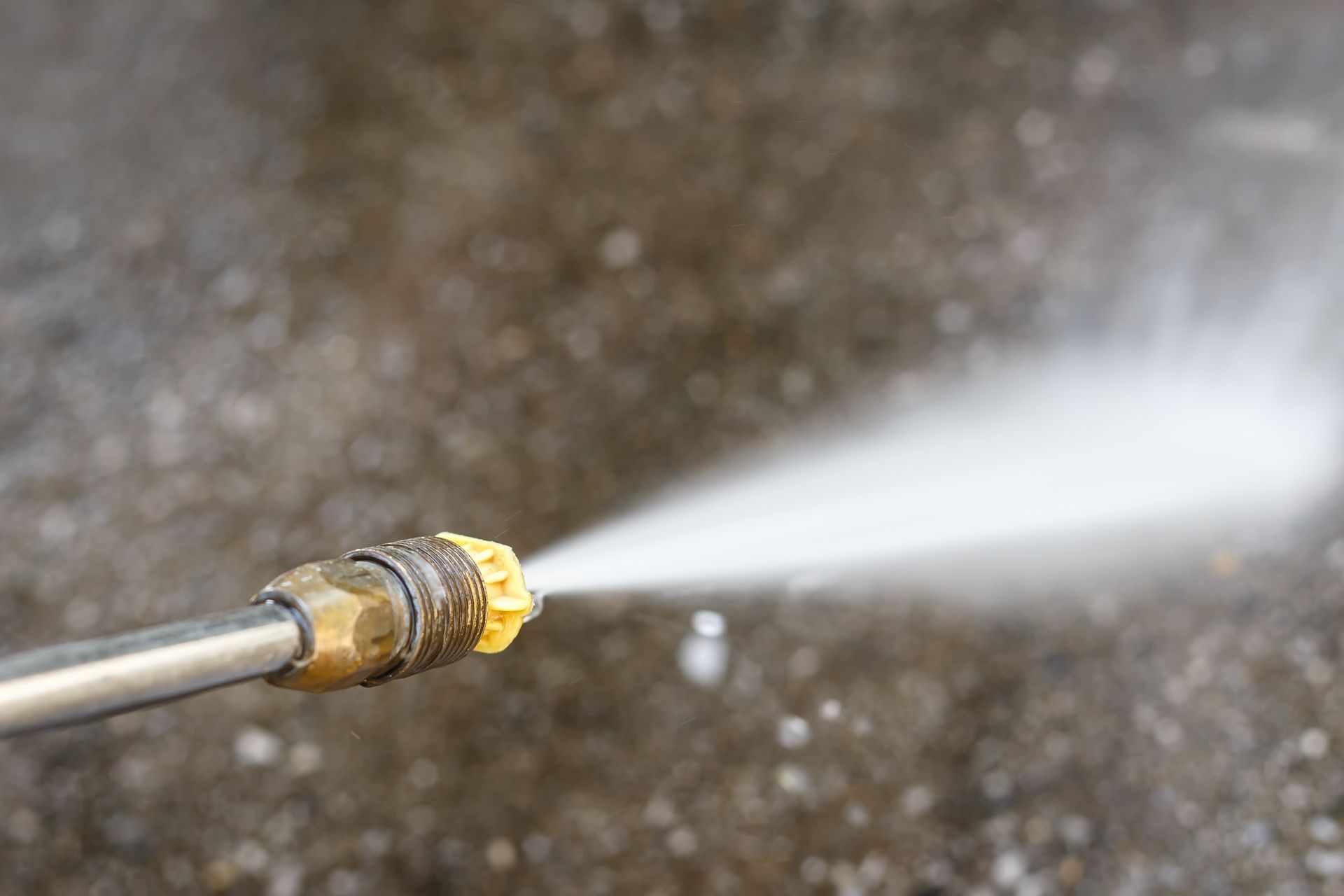 A pressure washer nozzle spraying a high-pressure stream of water onto a dark, textured surface.