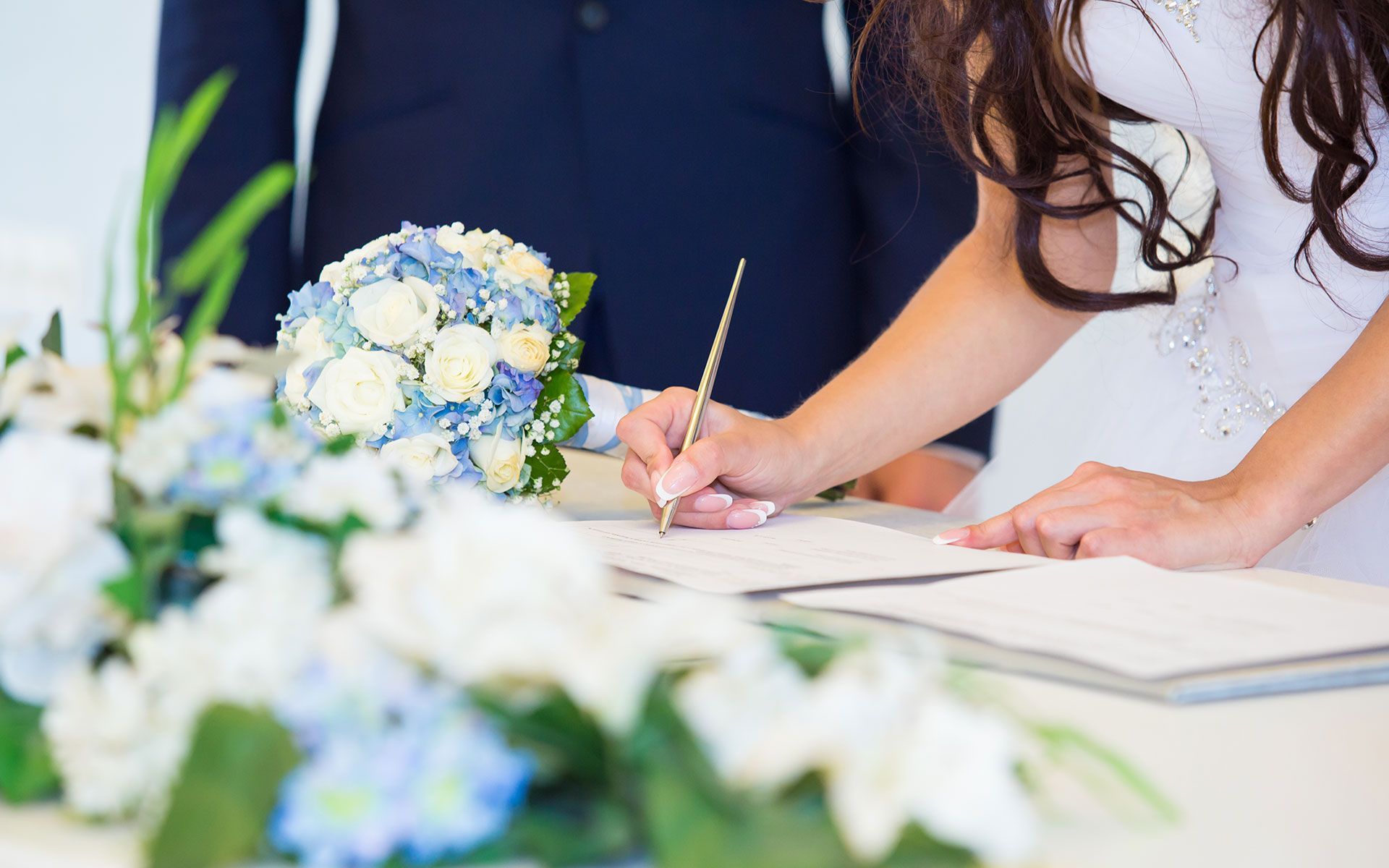 Bride signing marriage certificate, blue and white flower bouquet, man in navy suit.