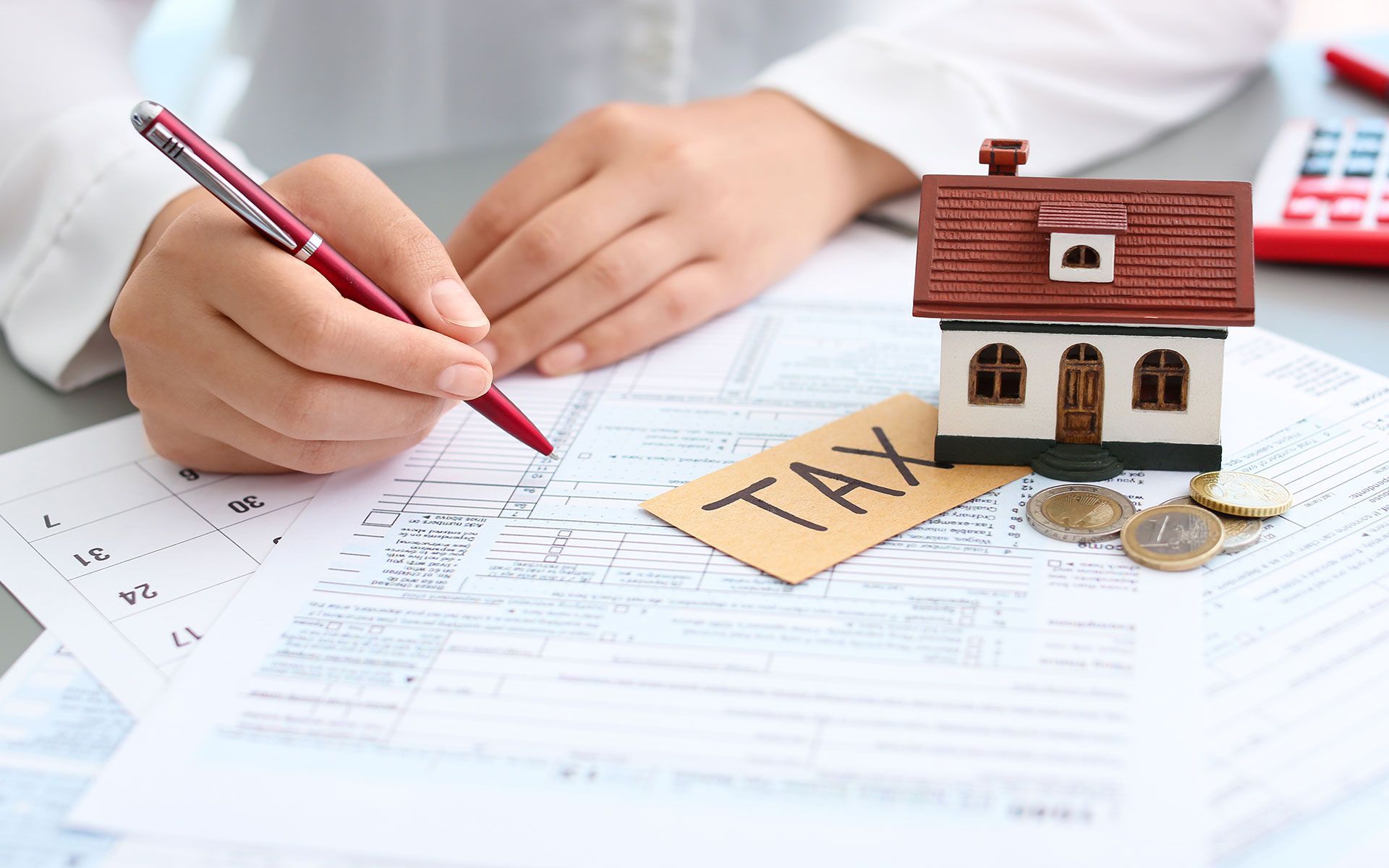 Person writing on tax forms with a toy house and coins, indicating property tax.