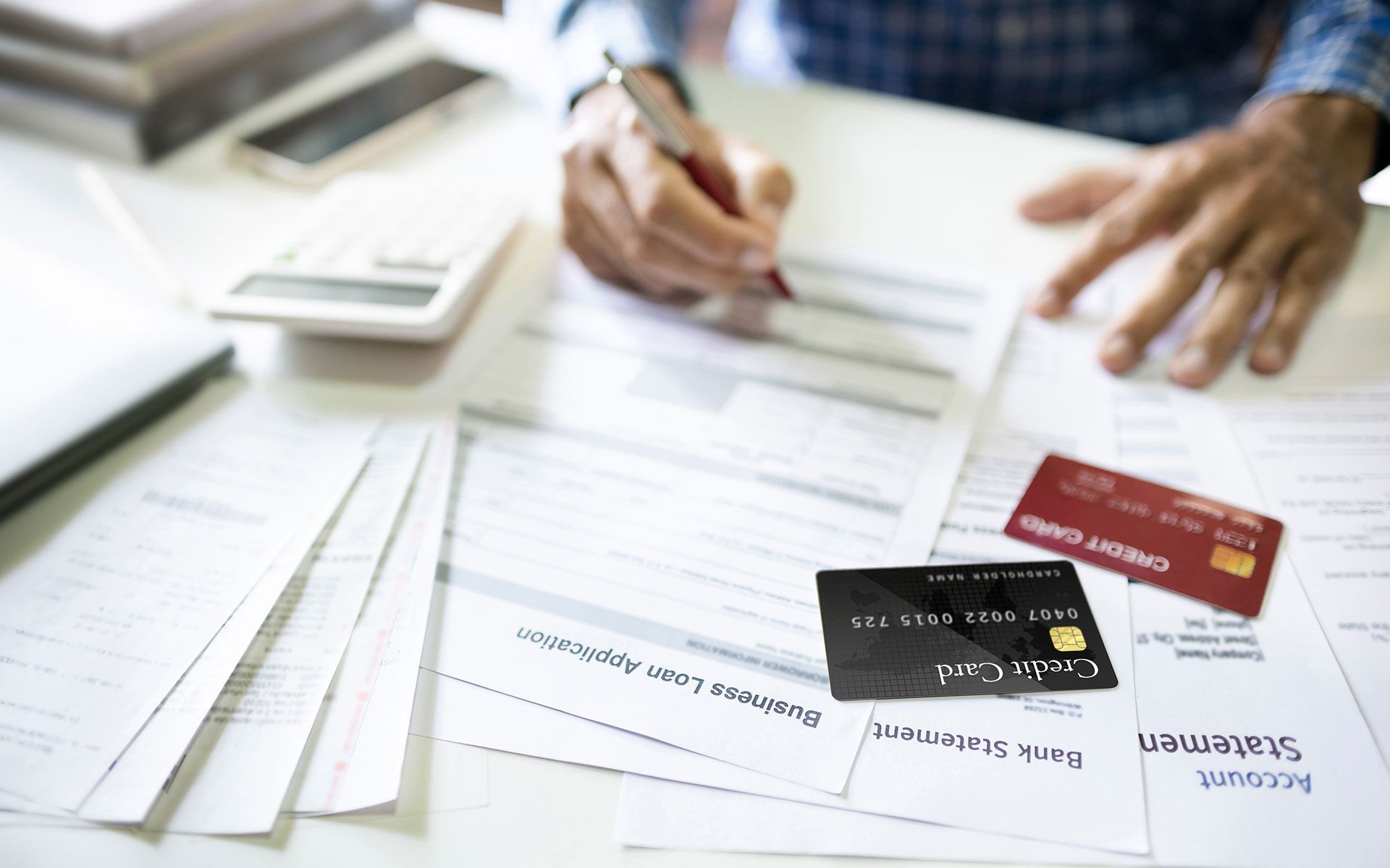Person filling out financial paperwork with credit cards and calculator on a white desk.