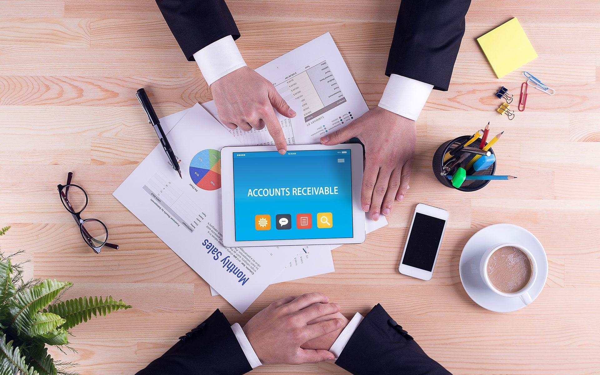 Two people at a desk review financial data on a tablet. Includes papers, a phone, and coffee.