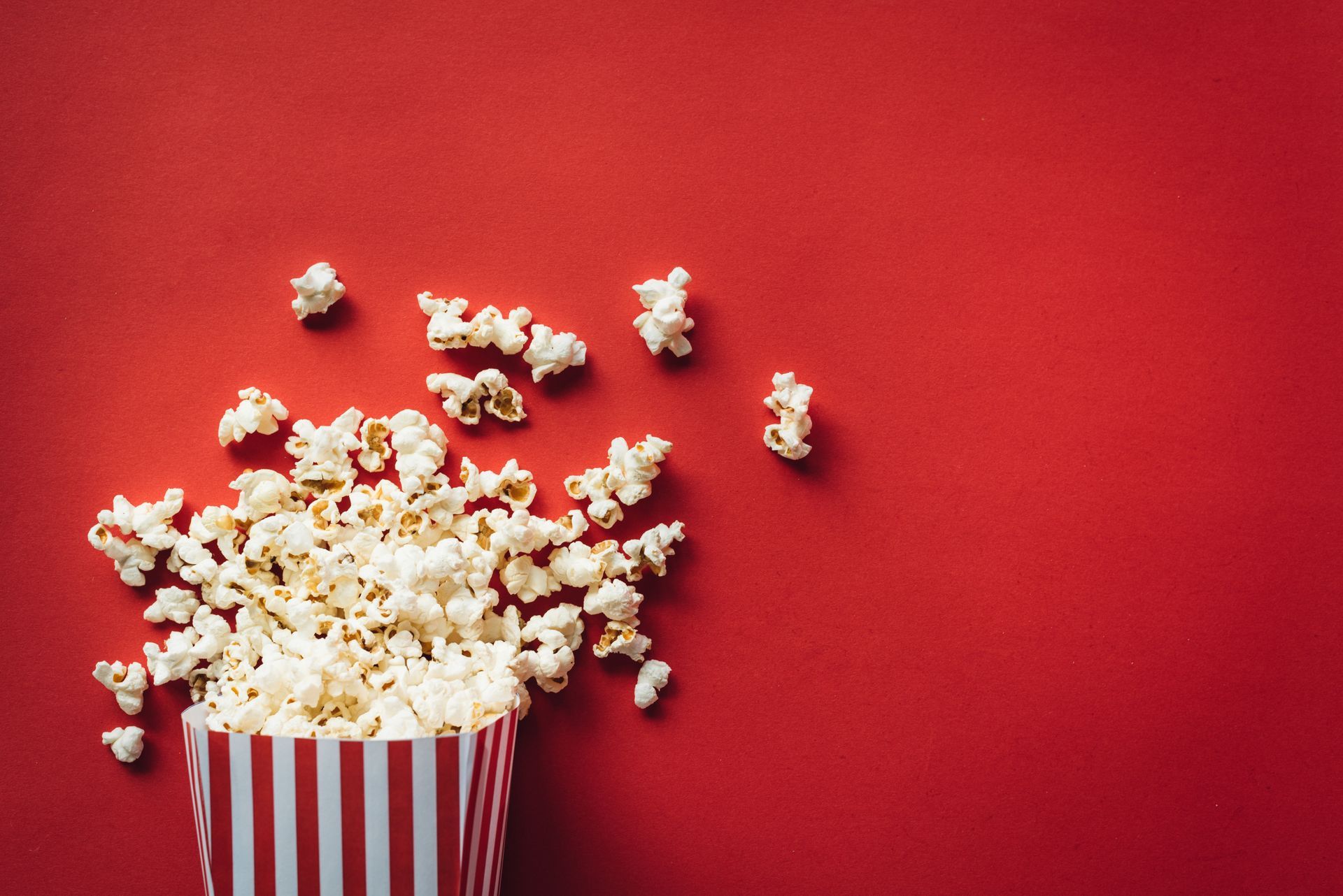 Popcorn is spilling out of a striped bucket on a red background.