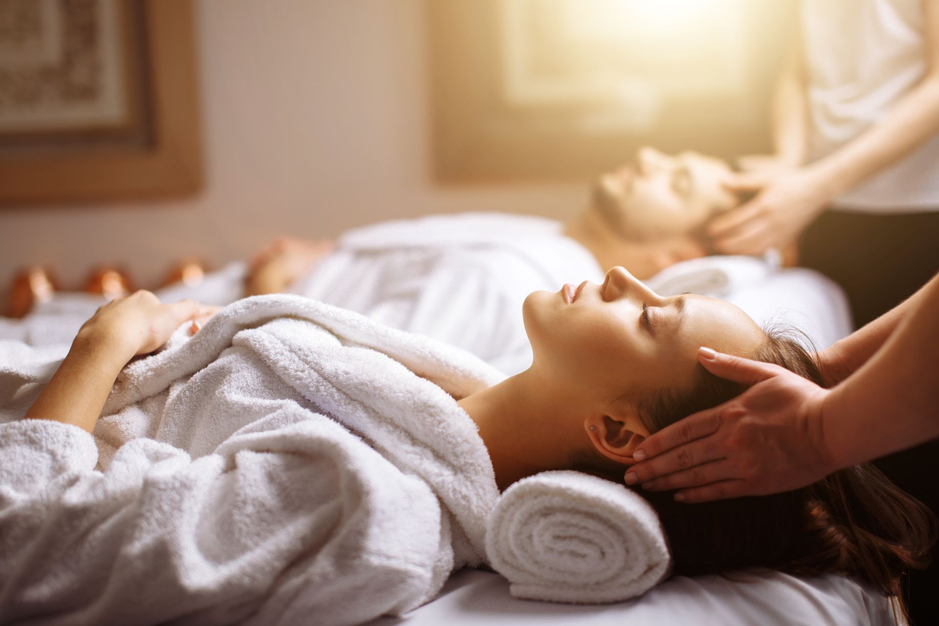 Couple receiving head massages at a spa. Woman and man in white robes, sunlight, serene expressions.