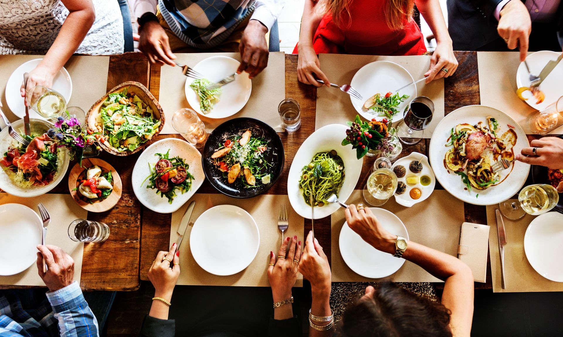 People at a wooden table enjoying a meal with various dishes, hands reaching for food.