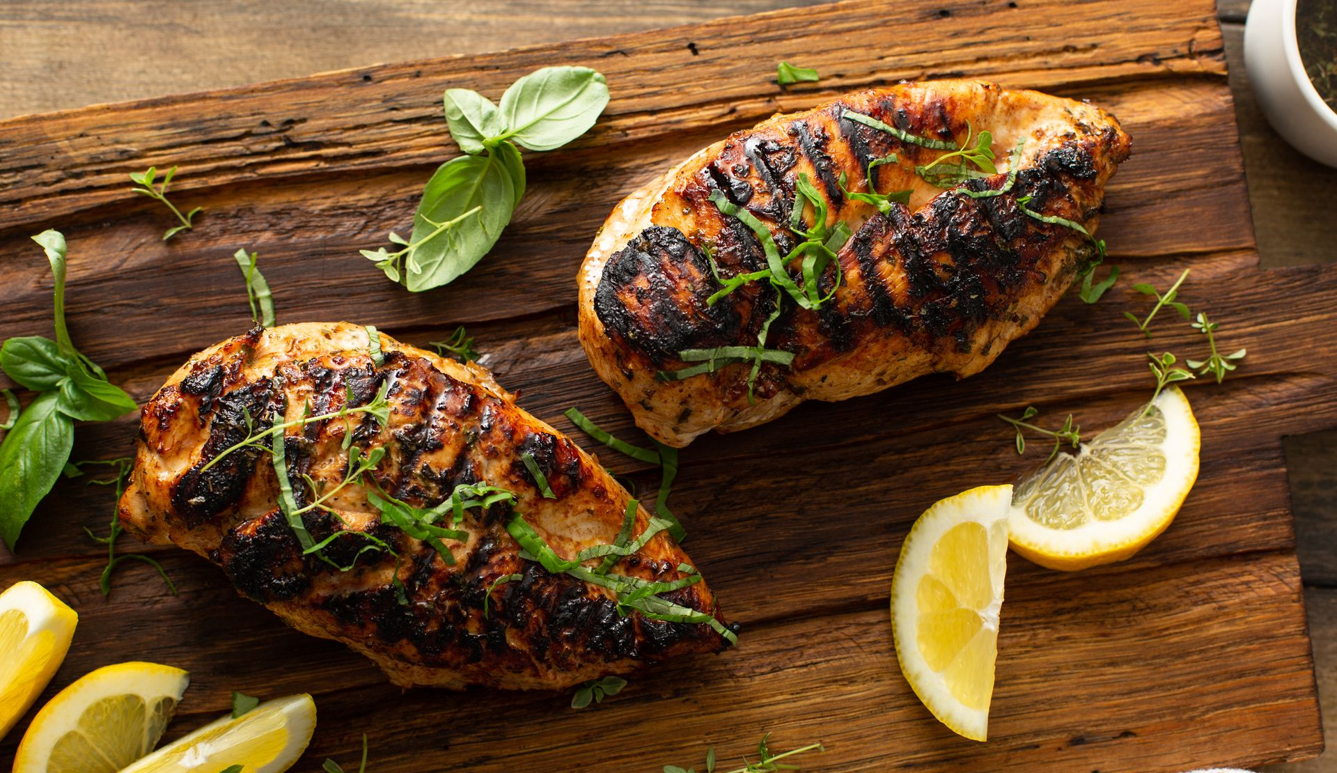 Grilled chicken breasts on a wooden board with lemon slices and herbs.