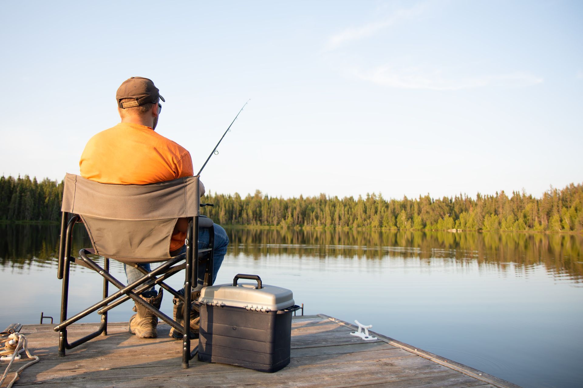 Man in an orange shirt fishing from a dock, facing a calm lake with a treeline in the background.