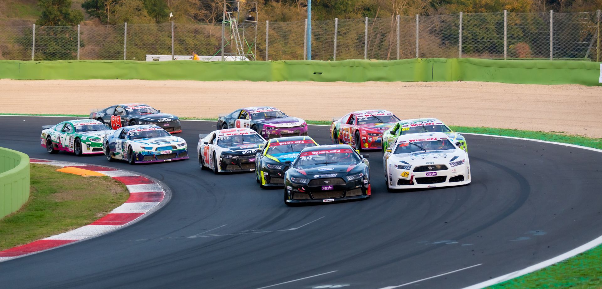 A pack of race cars navigates a corner on an asphalt track with a grass verge.