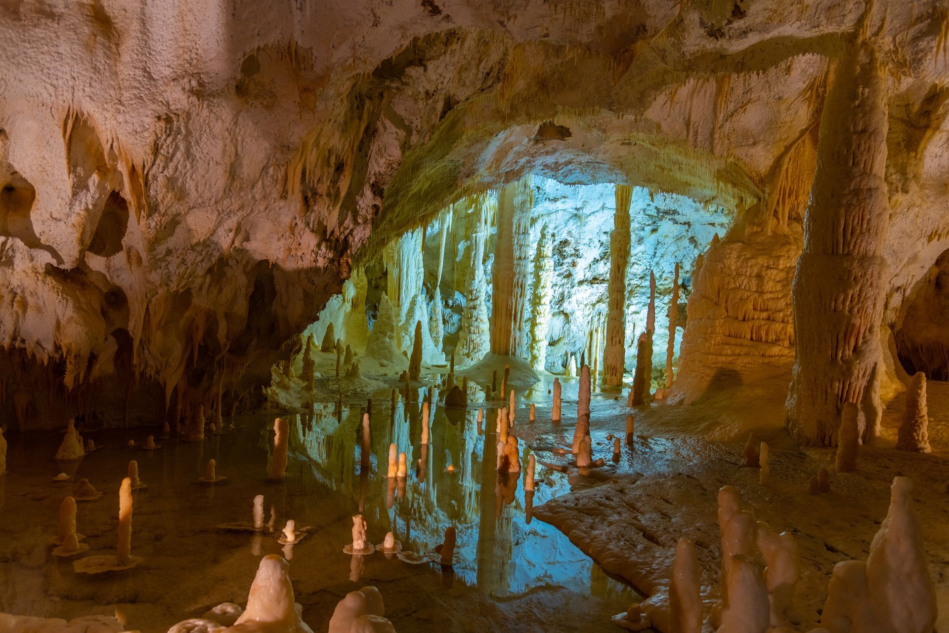 Cave interior with stalactites and stalagmites, reflecting in water, with a bright blue opening.