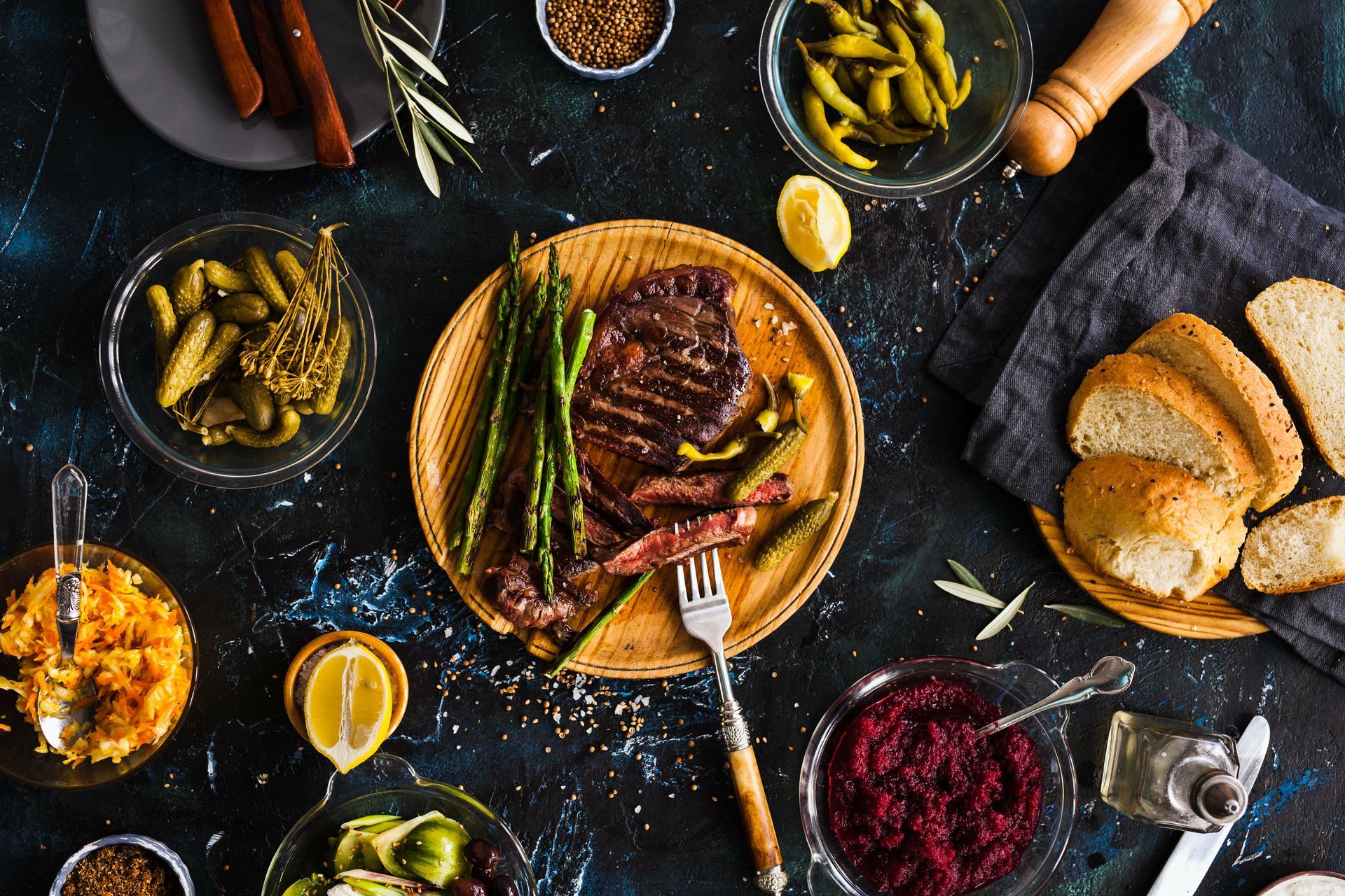 Overhead view of grilled steak and asparagus with various sides like pickles, bread, and sauces on a dark blue surface.