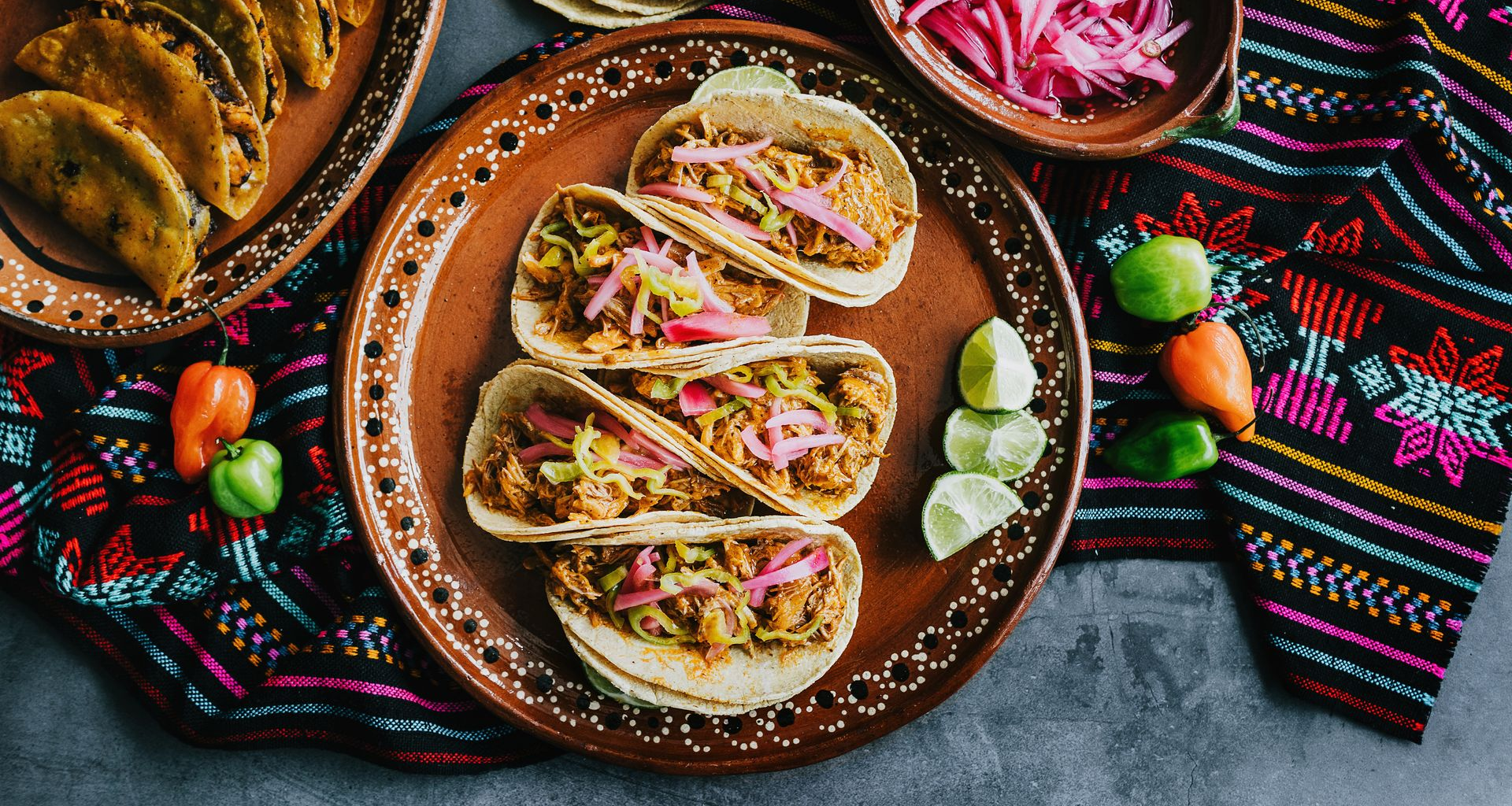 Tacos on brown plates with purple cabbage, limes, and peppers on colorful cloth.