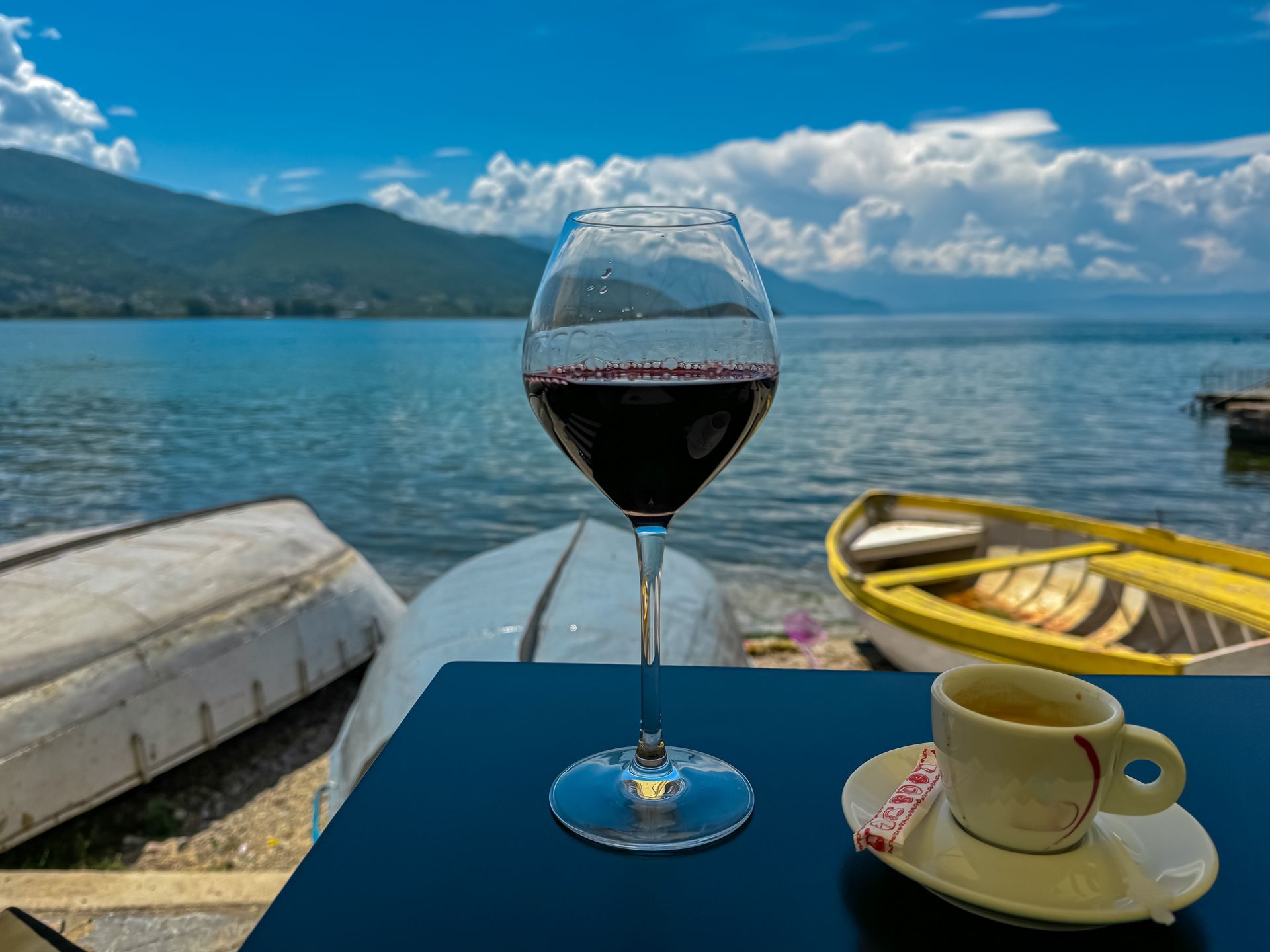 Wine glass and espresso cup on a table overlooking a lake and boats, mountains in the background, sunny day.