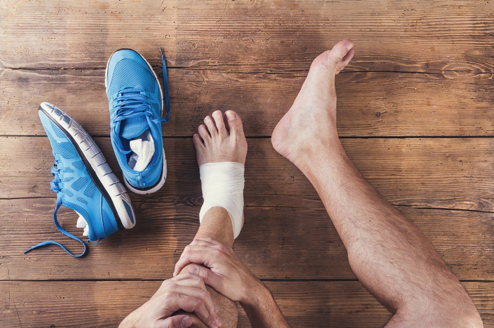 Person's injured ankle wrapped in bandage, next to blue running shoes on a wooden floor.