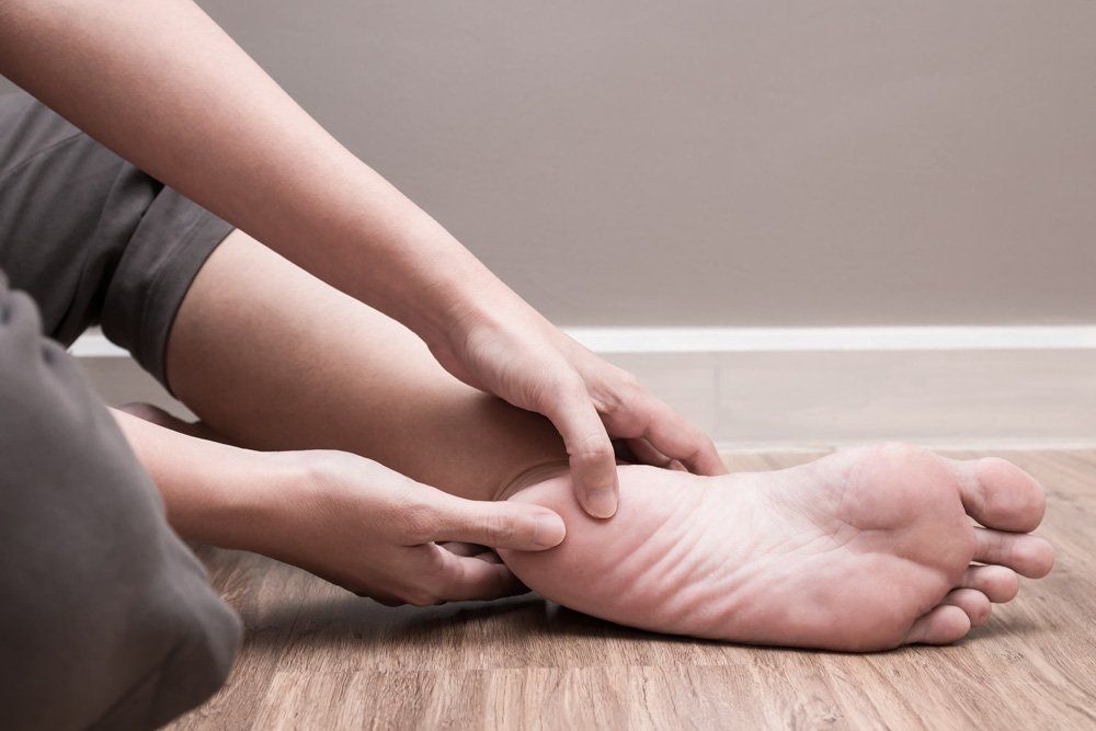Person touching their foot, possibly in pain, indoors on wooden floor.