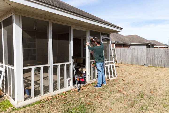 Person standing on a porch adjusting a white screen frame beside patio furniture and a yard fence