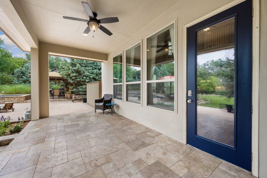 Covered patio with stone floor, ceiling fan, blue door, and large windows overlooking a backyard.