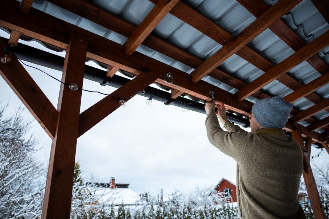Person fastening corrugated roofing under a wooden shelter in a snowy outdoor setting