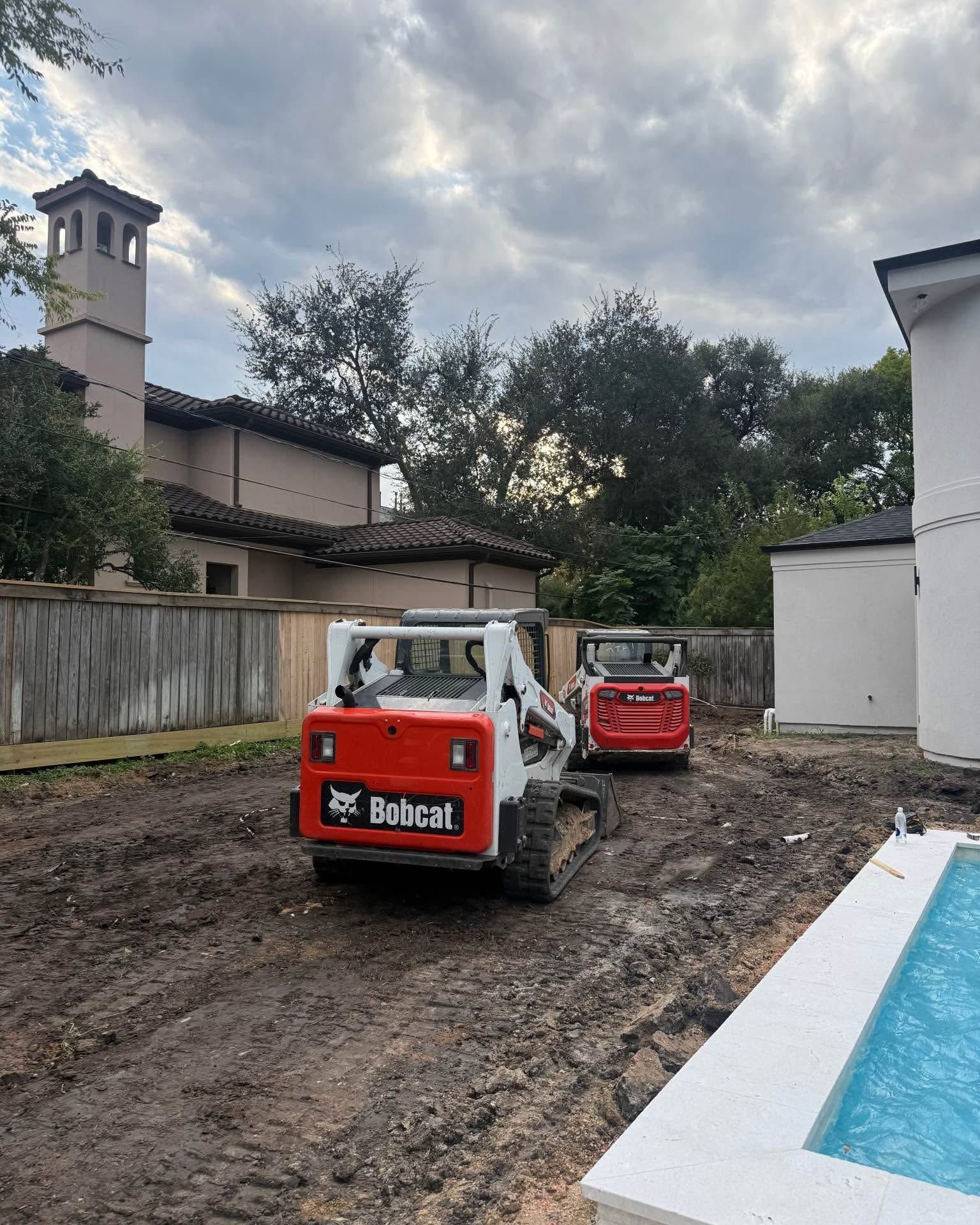 Two Bobcat track loaders parked on a muddy residential lot next to a swimming pool.
