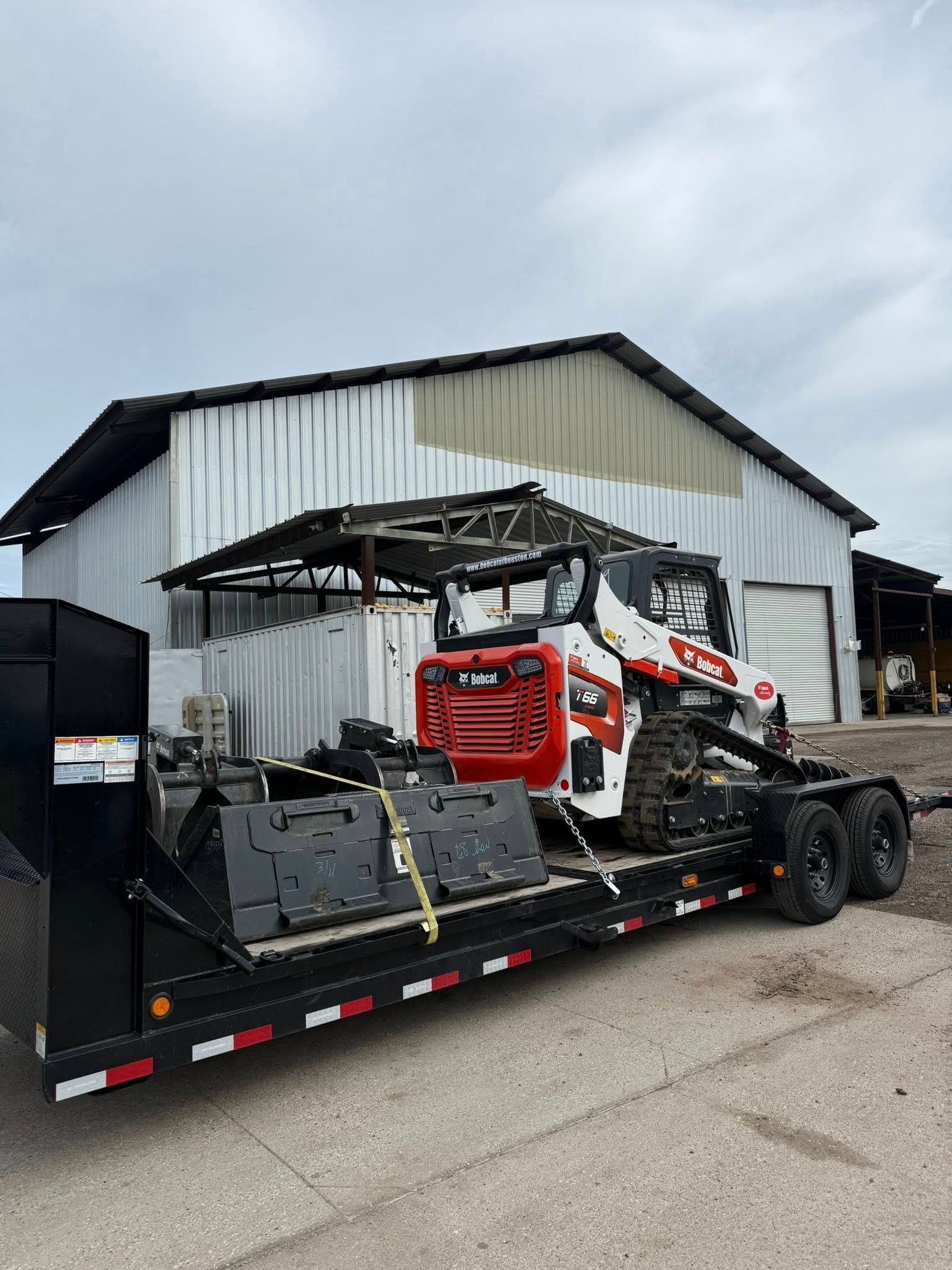 A white and red Bobcat skid-steer loader sits secured on a flatbed trailer in front of a metal warehouse.