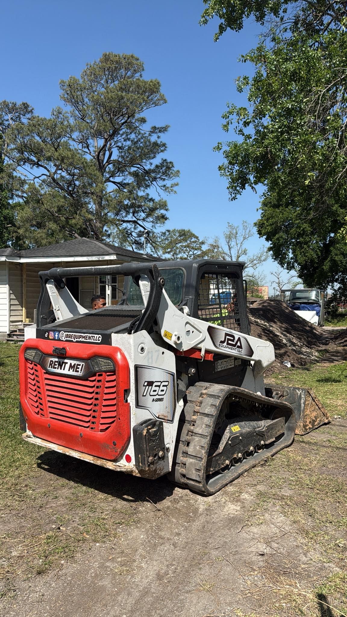 A white and red compact track loader parked on a grassy lot in front of a house on a sunny day.