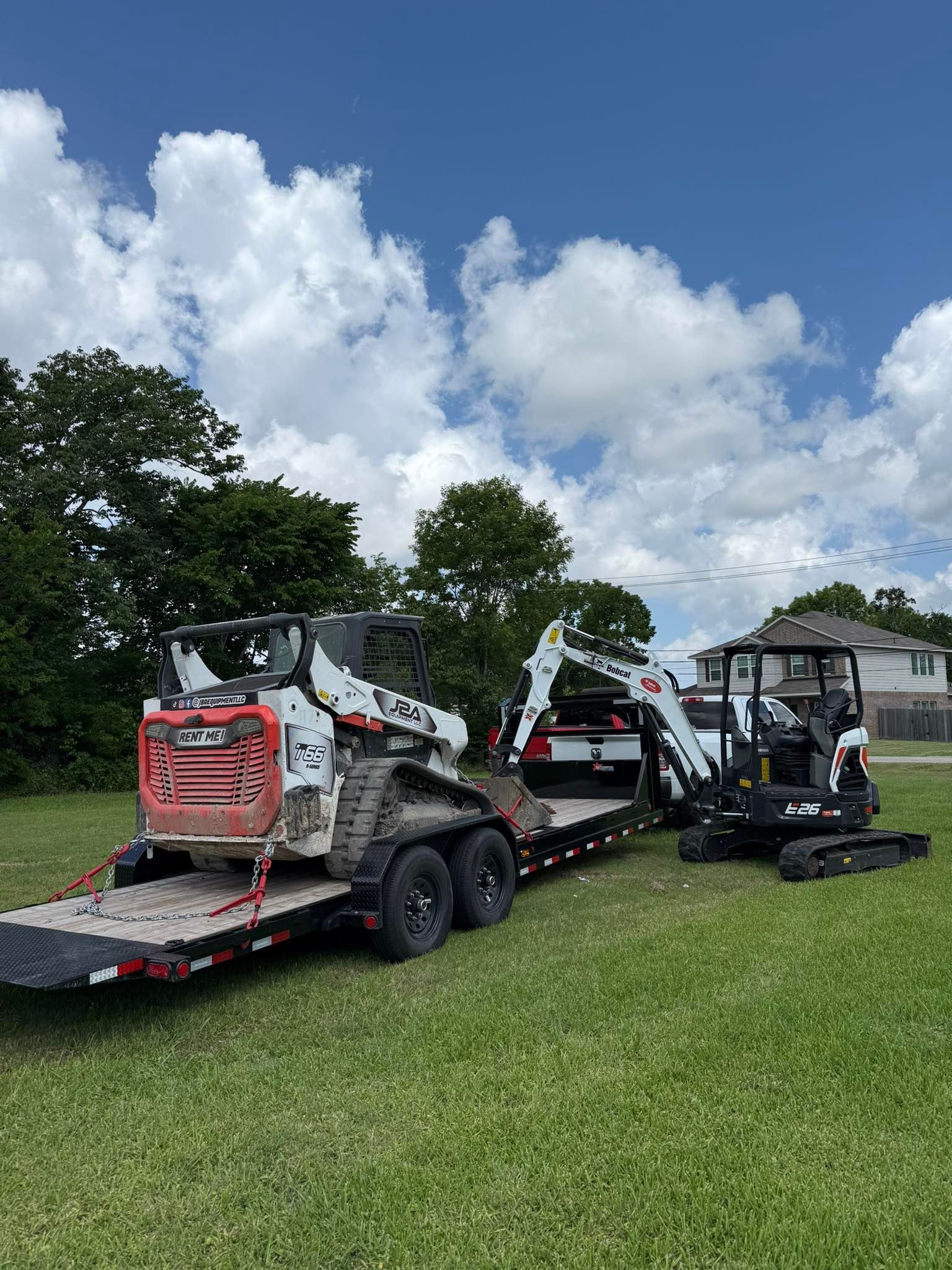 A white and red skid-steer loader sits on a flatbed trailer being towed by a truck, with a mini excavator parked nearby.