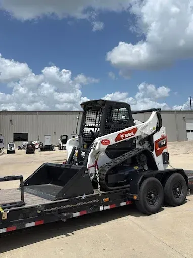 A white and black Bobcat compact track loader loaded on a flatbed trailer in a paved lot under a blue, cloudy sky.