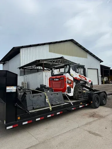 A white and red Bobcat track loader sits secured on a flatbed trailer in front of a metal warehouse.