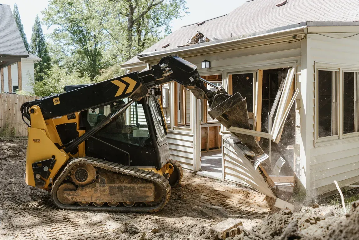 A yellow and black skid-steer loader tears down the siding and frame of a small white house in a backyard setting.