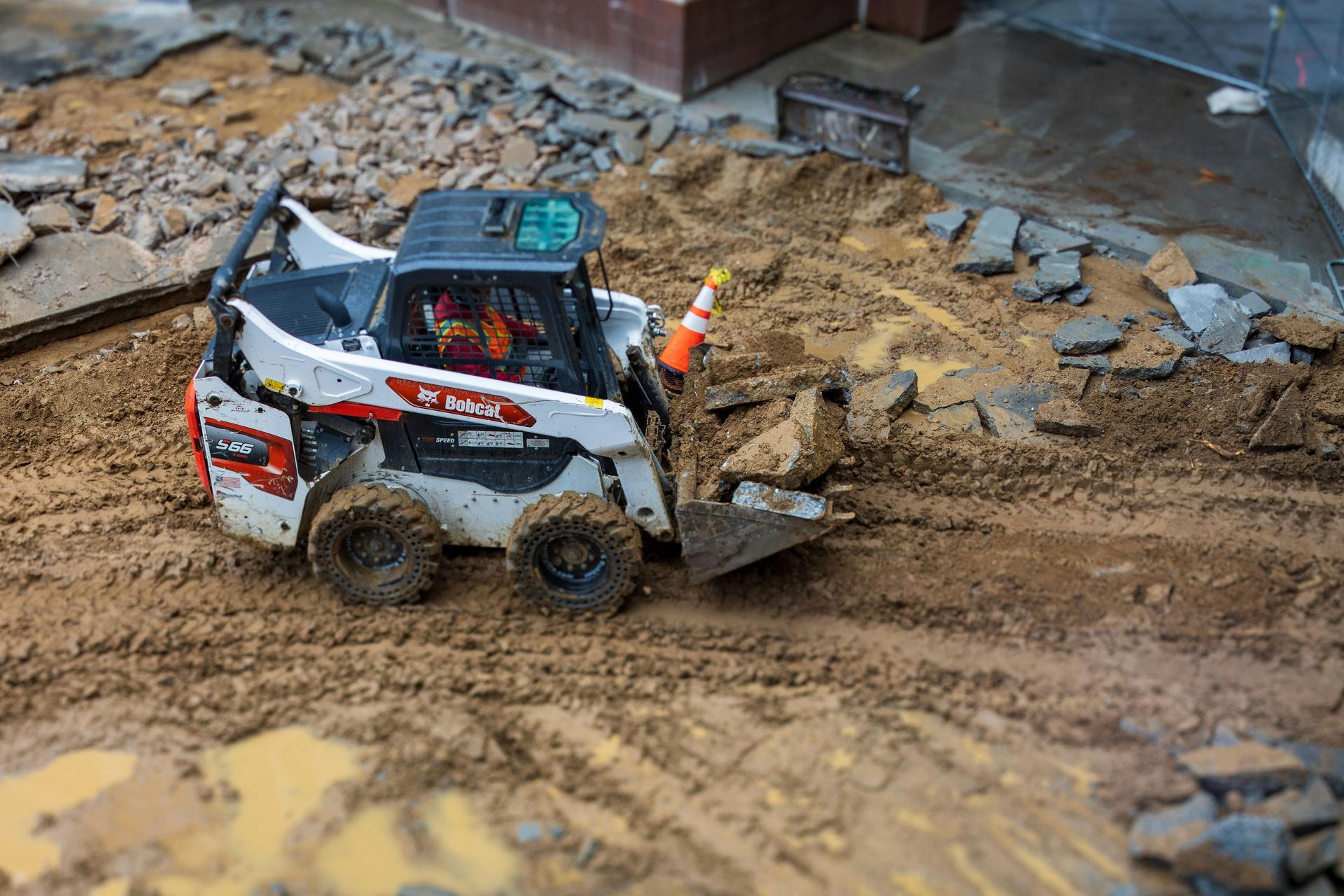 A Bobcat skid-steer loader clears dirt and debris on a muddy construction site next to a small traffic cone.