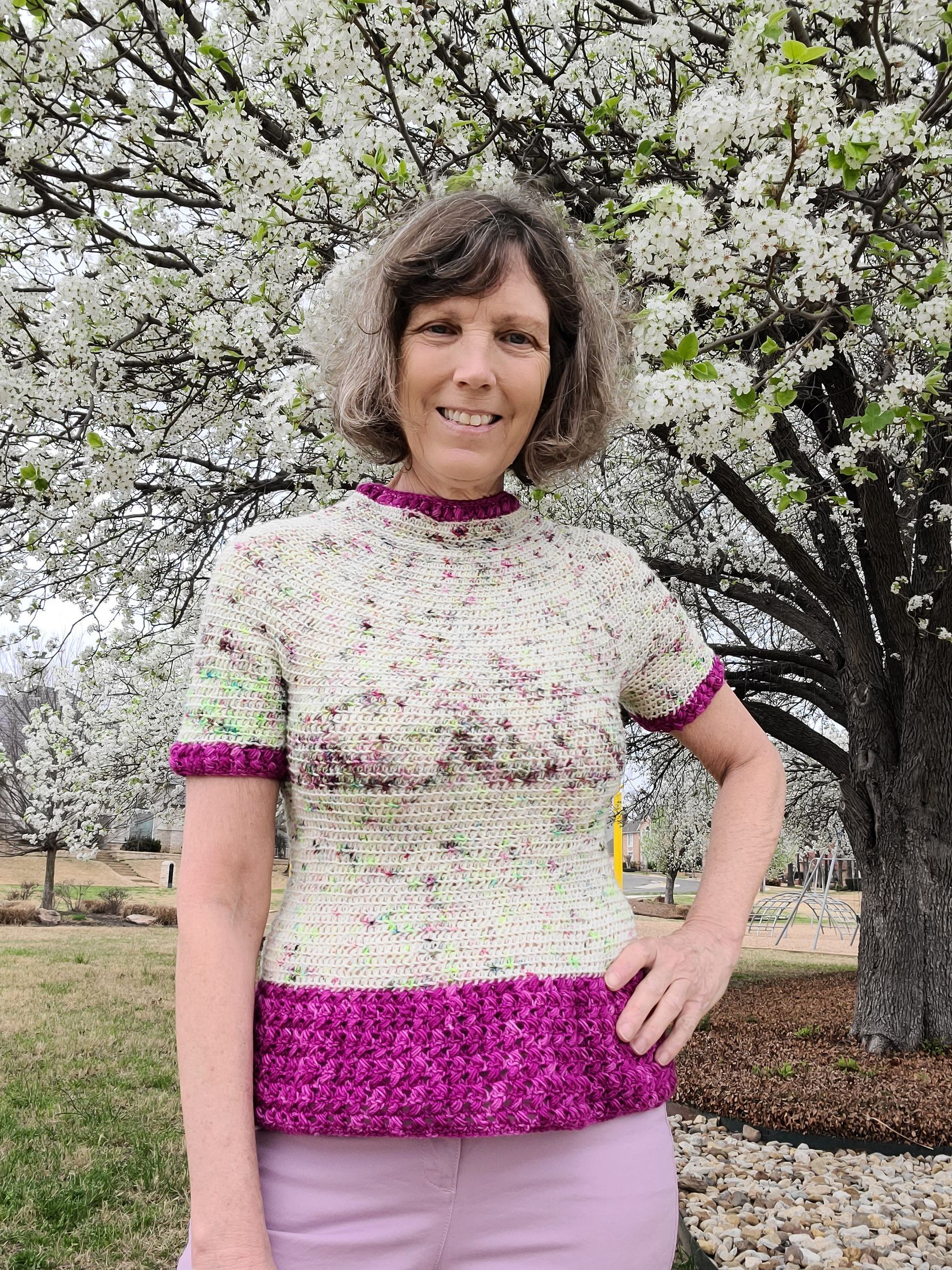 Woman in a hand-knitted sweater, with a blooming tree background. She is smiling, standing outside.