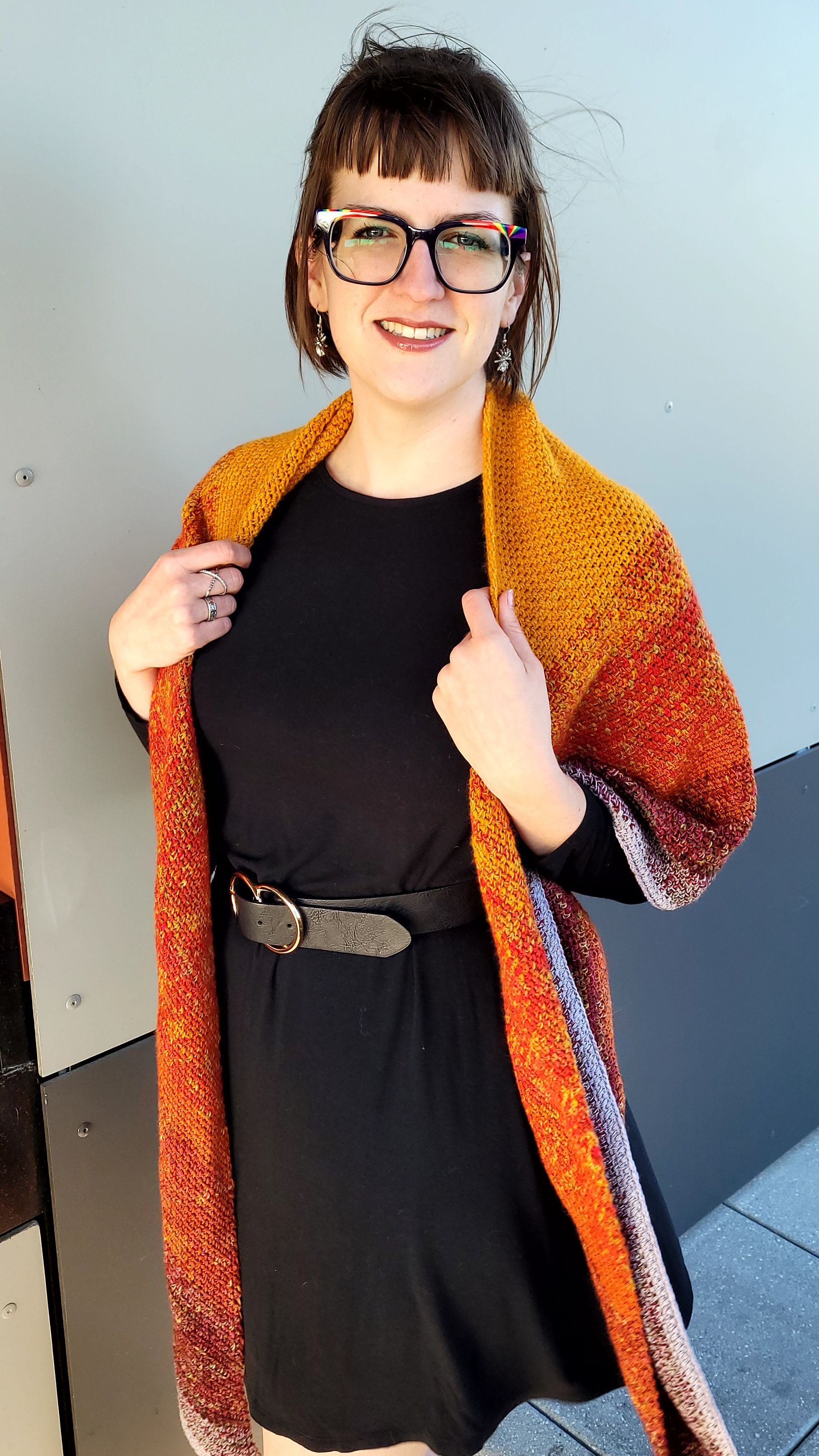 Woman in glasses, black dress, and orange shawl smiles, standing outdoors.