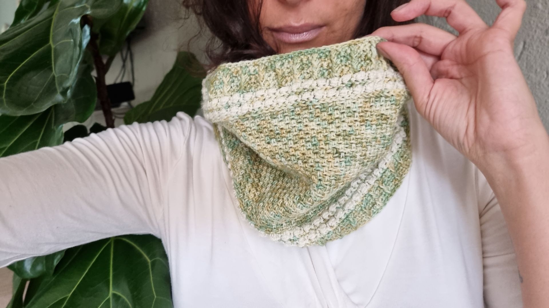 Woman holding a green and white knit cowl, indoors near a leafy plant.