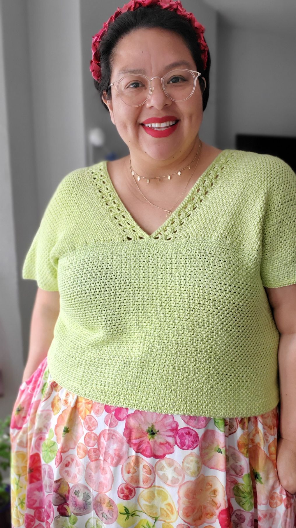 Woman in green knit shirt and floral skirt smiling, wearing glasses and a headband.