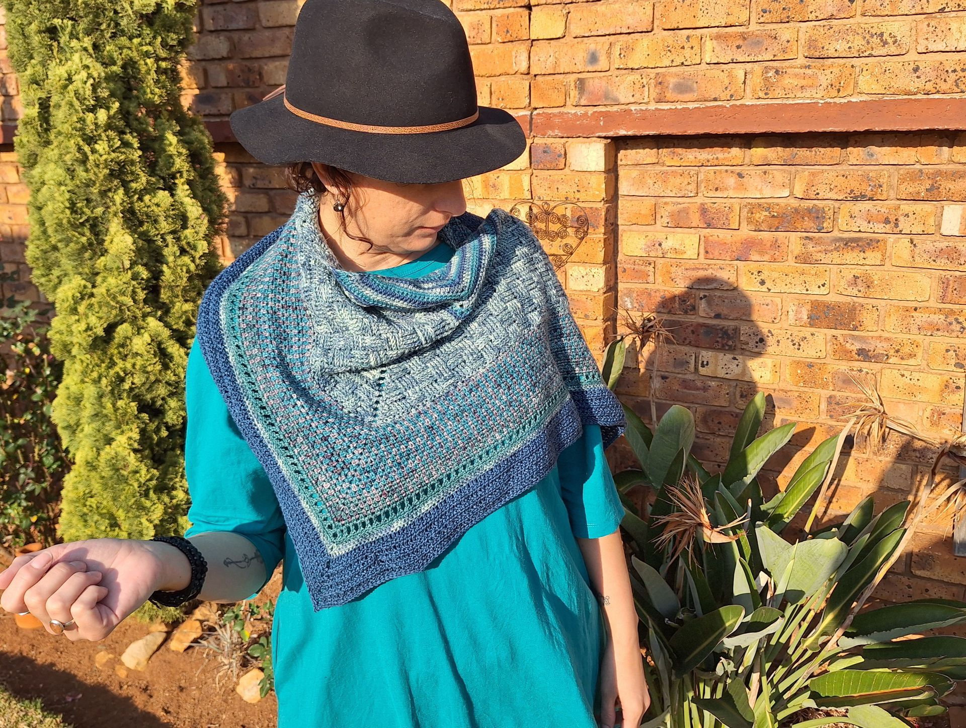 Woman wearing blue shawl and hat, outdoors near brick wall and plants.