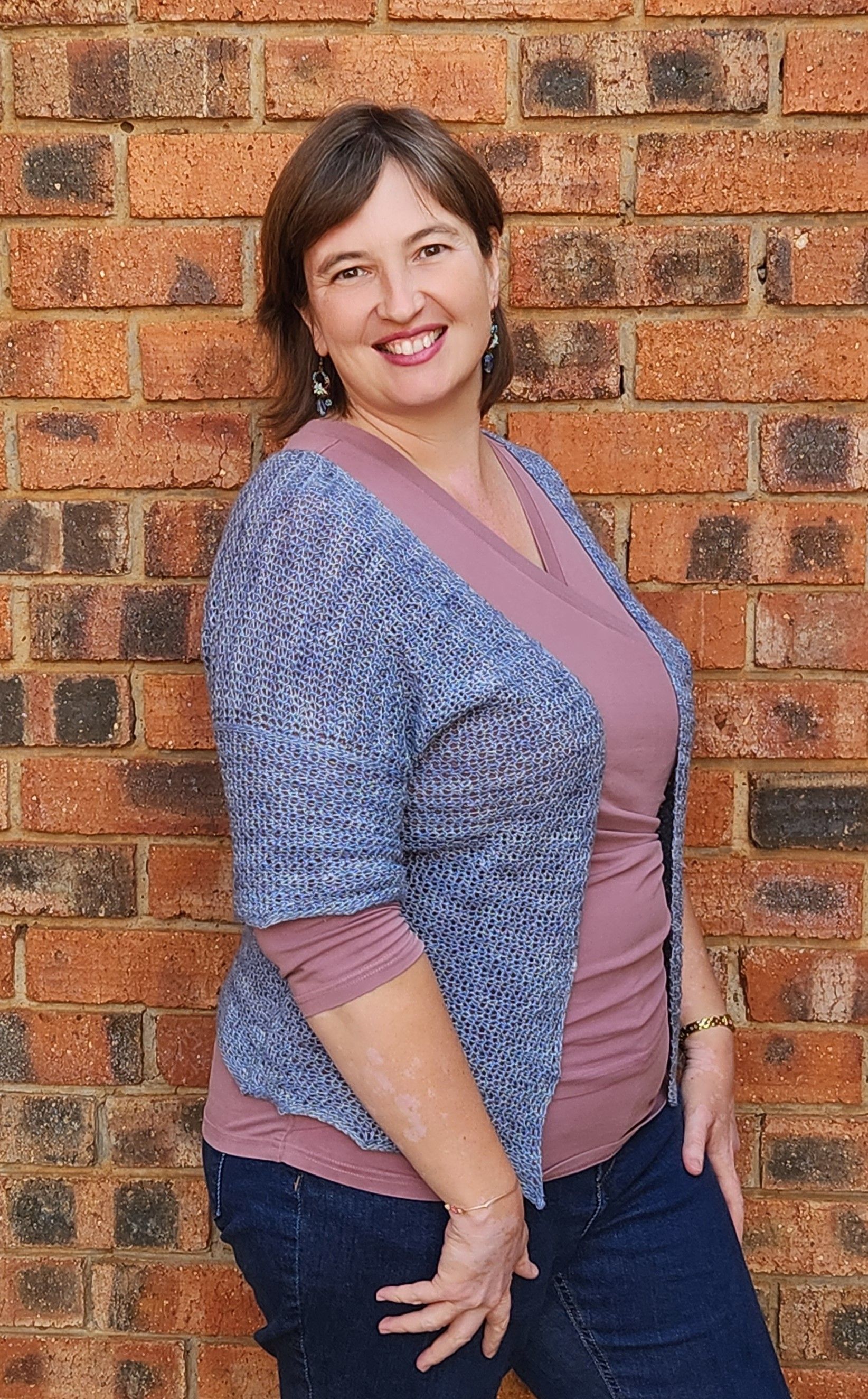 Woman smiles, leaning against brick wall. She wears pink top, blue cardigan, and jeans.