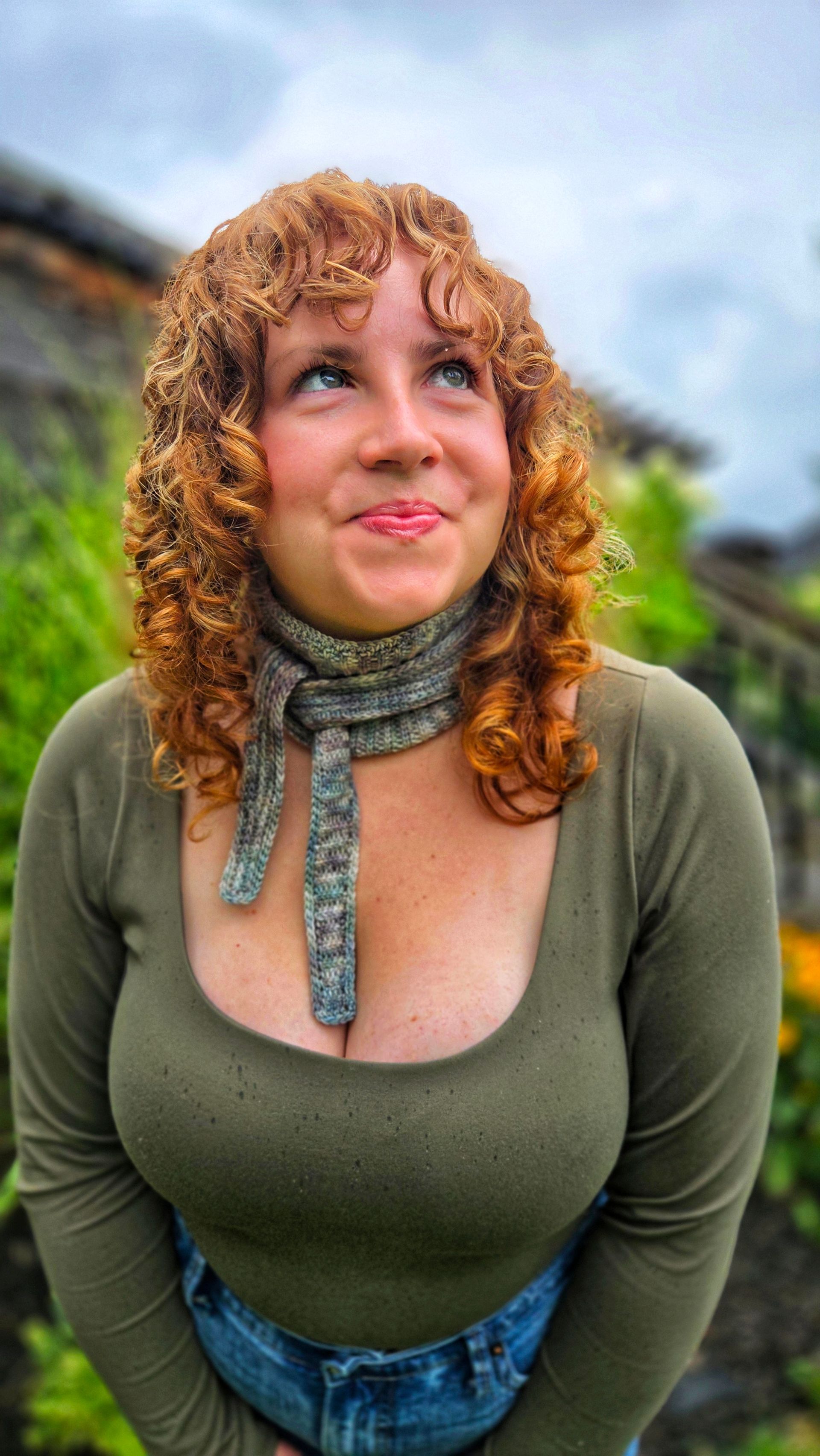 Woman with curly red hair looking up, wearing olive green top and scarf, smiling. Outdoors.