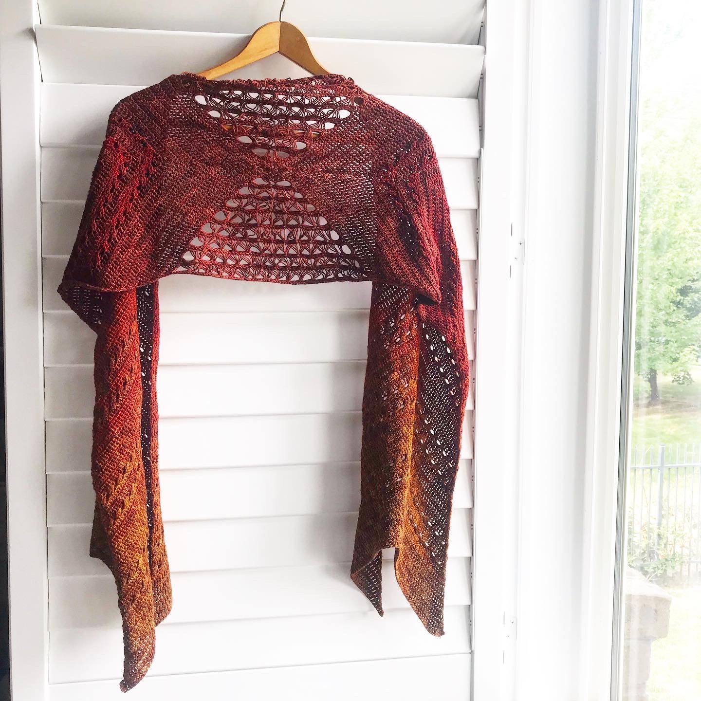 A crocheted shawl with a gradient of red, orange, and brown hanging on a wooden hanger in front of white shutters.
