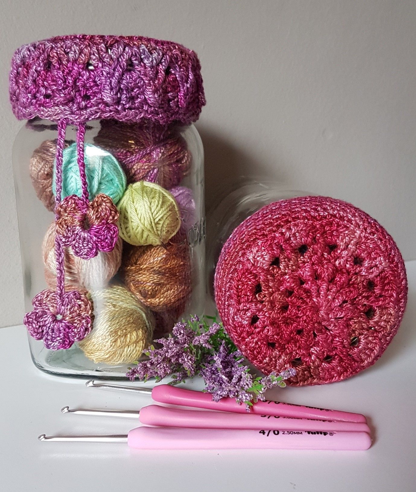 Crochet supplies: jar of yarn with a crocheted lid, crochet hook, and a crocheted ball in pink tones.