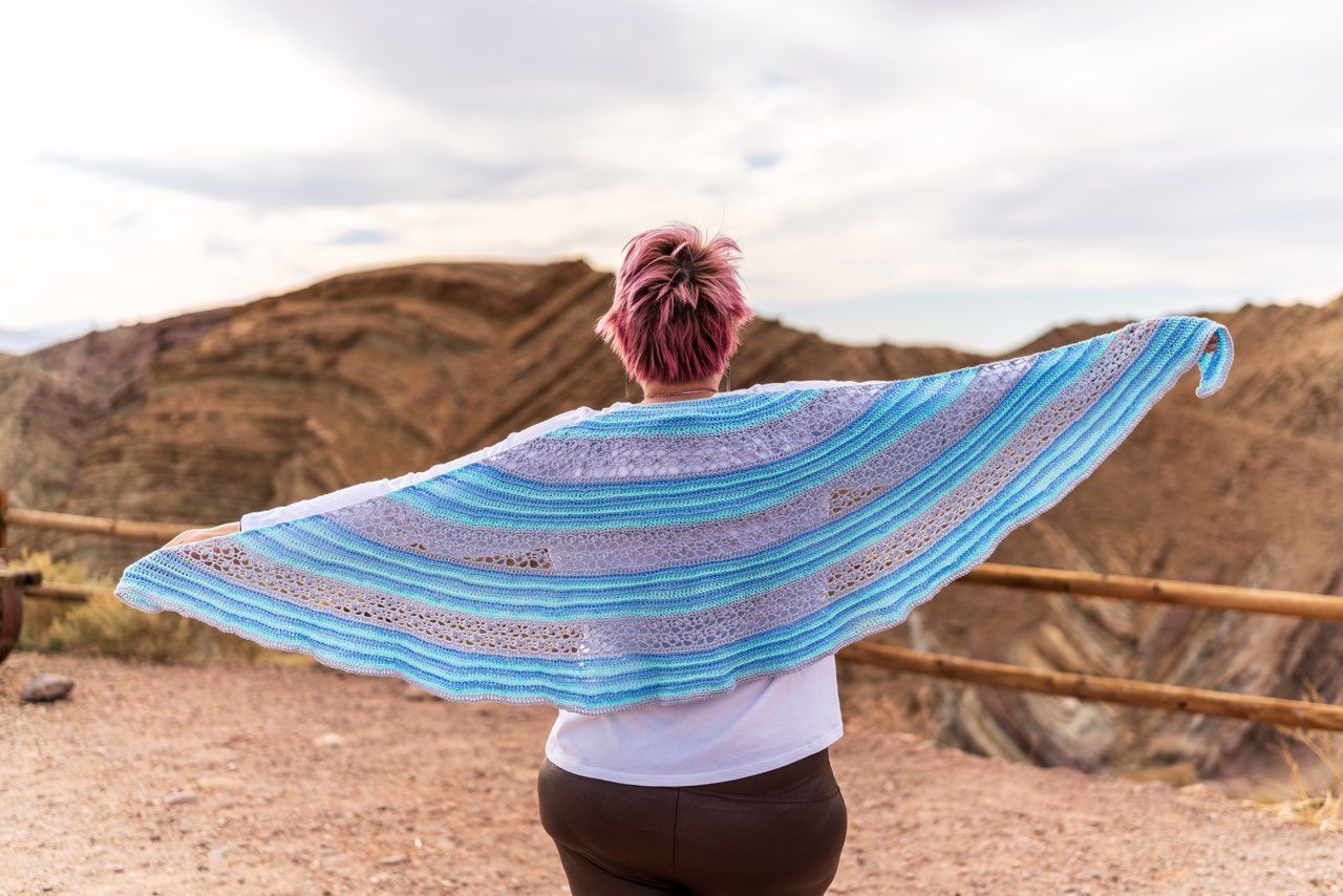 Woman with pink hair, holding a blue and gray striped shawl open, overlooking a mountain range.