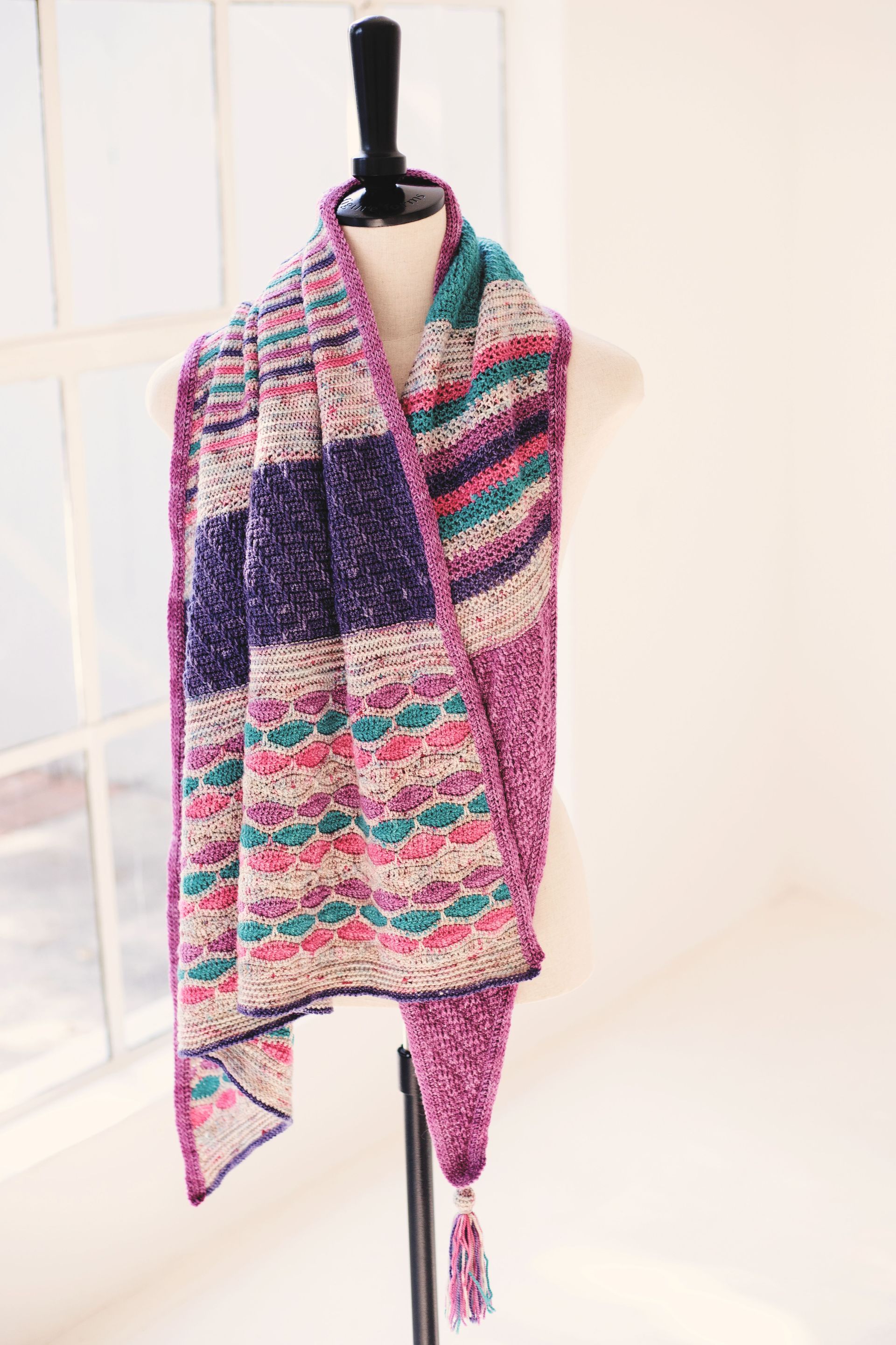 Crocheted scarf with purple, pink, and teal stripes, hanging on a mannequin.
