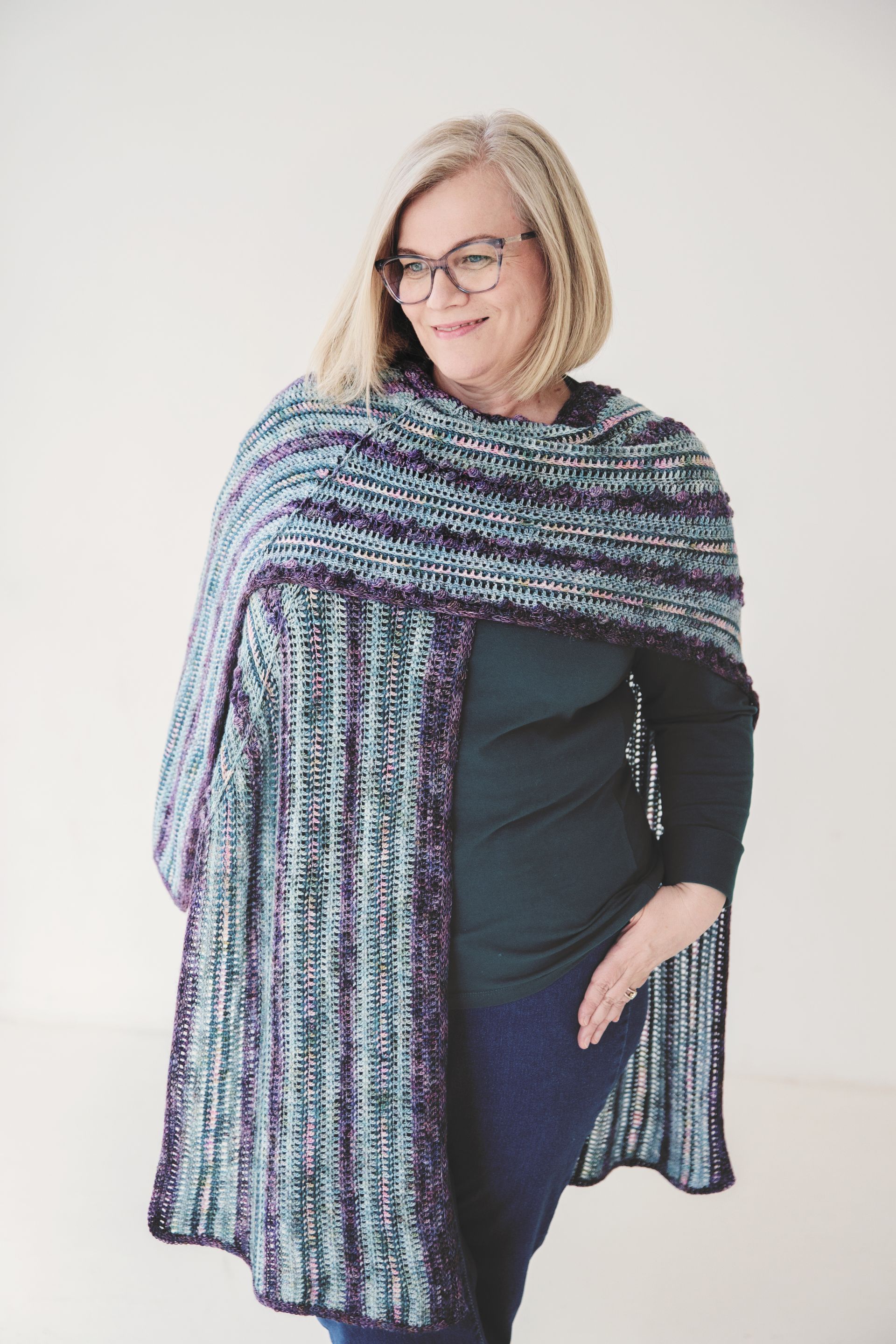 Woman in glasses, wearing a blue, purple, and green knit shawl. Standing against a white background.