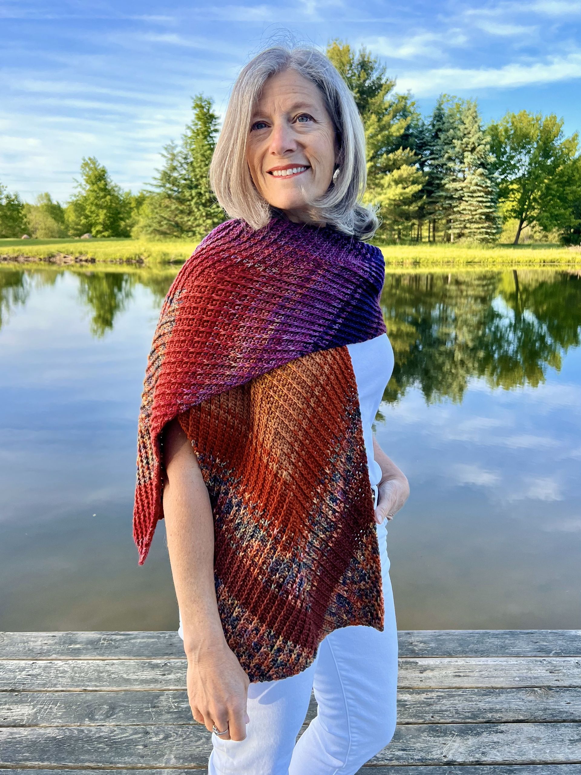 Woman wearing a gradient shawl, standing on a dock, with water and trees in background.
