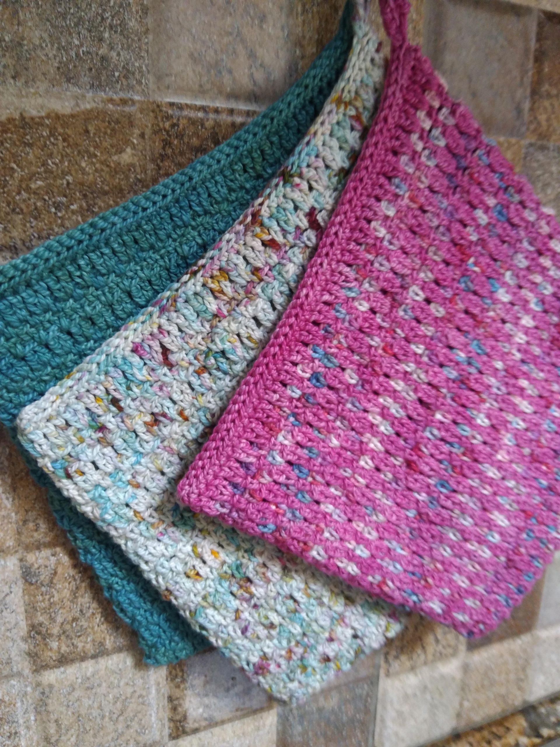 Three colorful crocheted potholders hanging on a textured wall: teal, white with speckles, and pink.