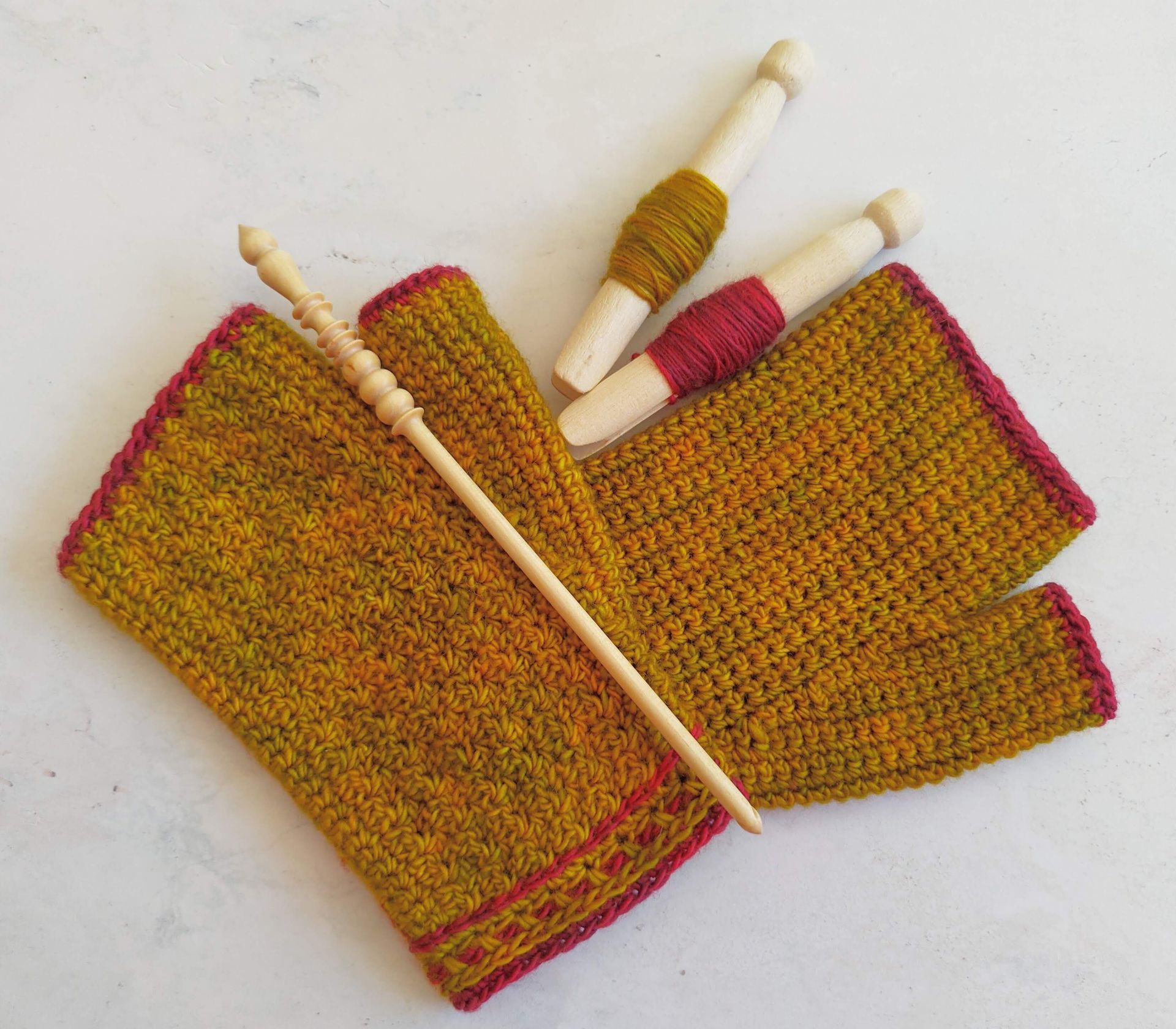 Crocheted mustard-colored hand warmers with red trim, wooden crochet tools on a white surface.