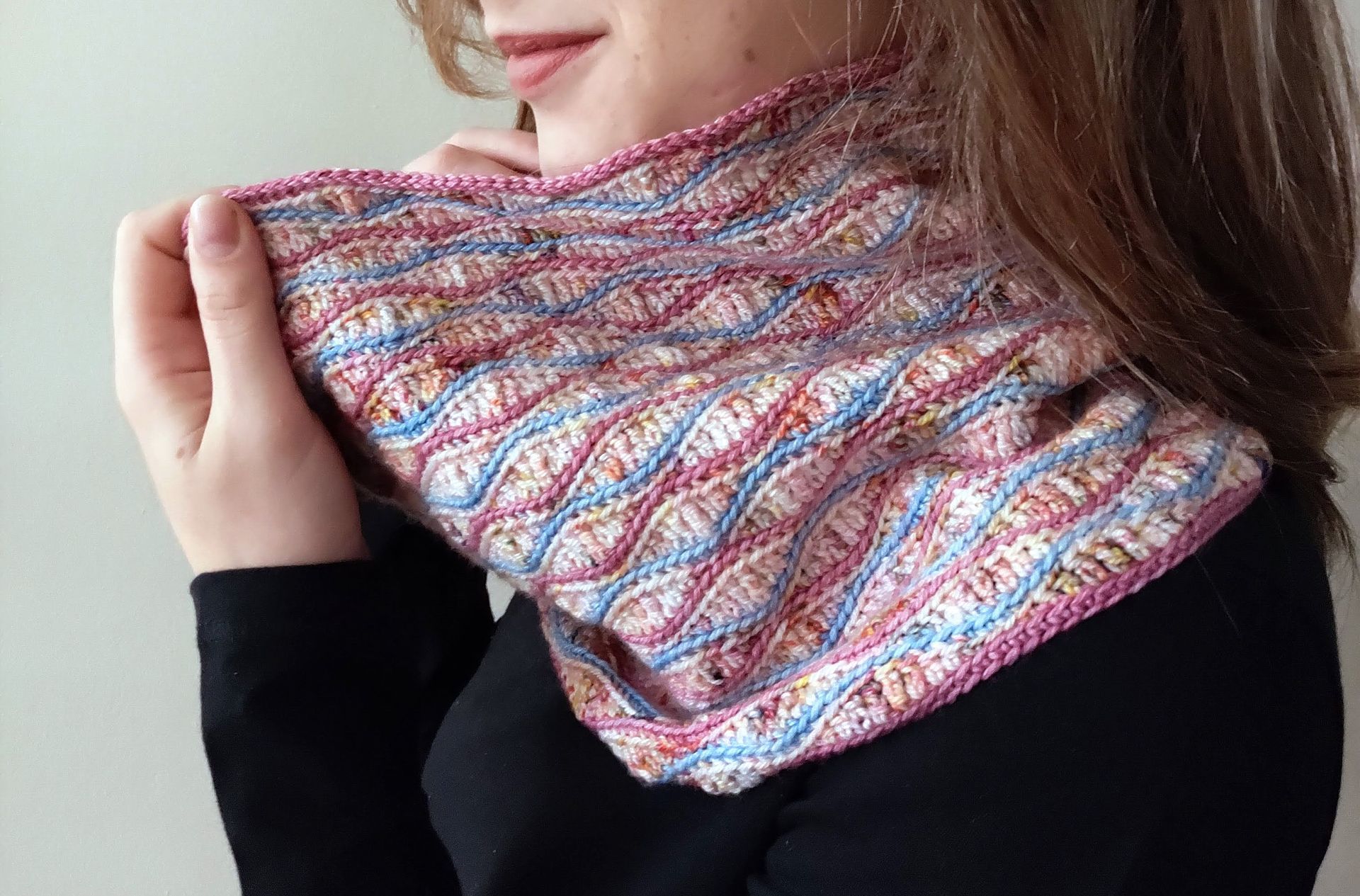Woman holding a colorful knitted cowl scarf. Pink, blue, and tan pattern. Black shirt.