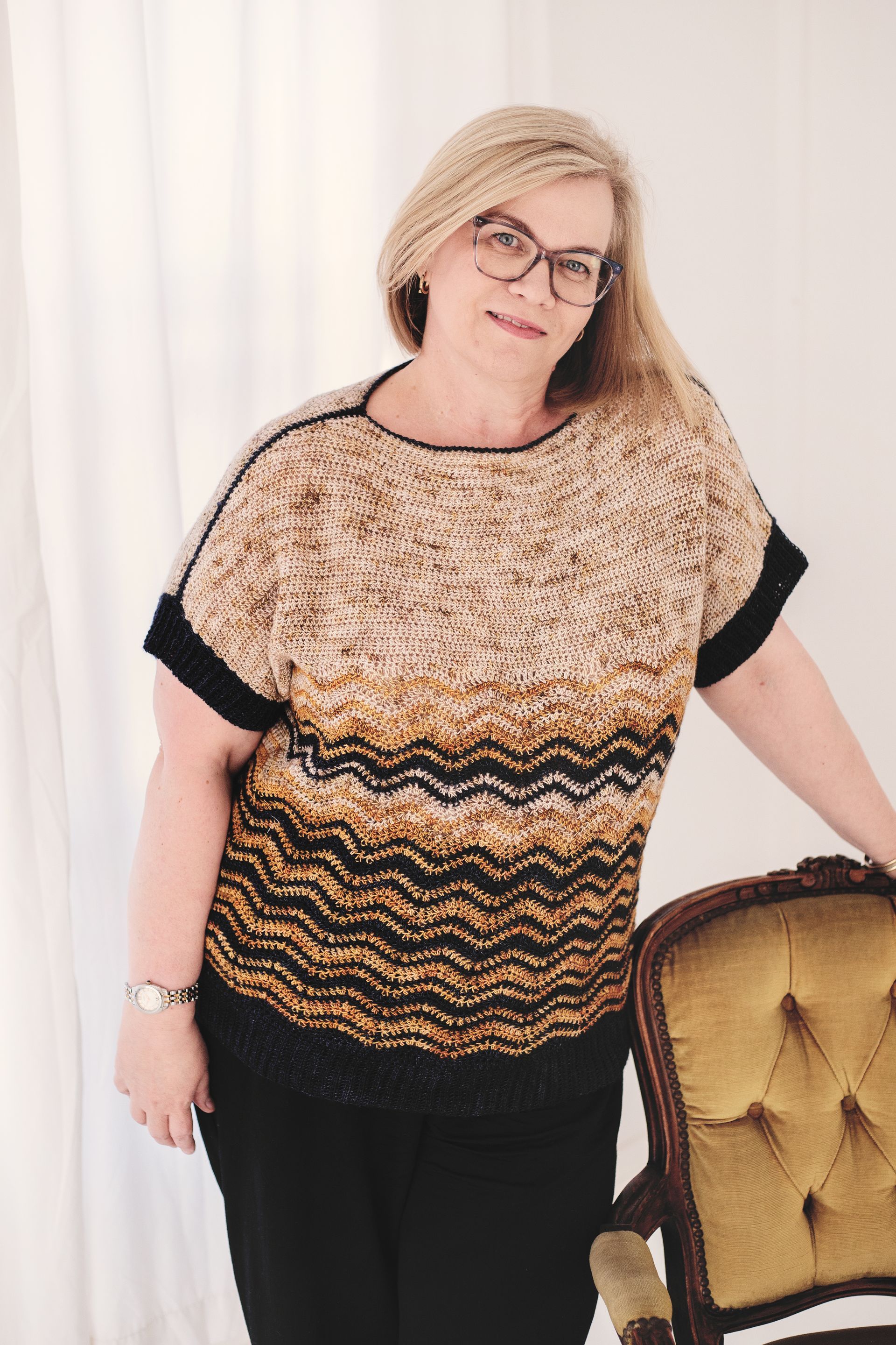 Woman wearing a patterned sweater, leaning against a chair, smiling; bright room.