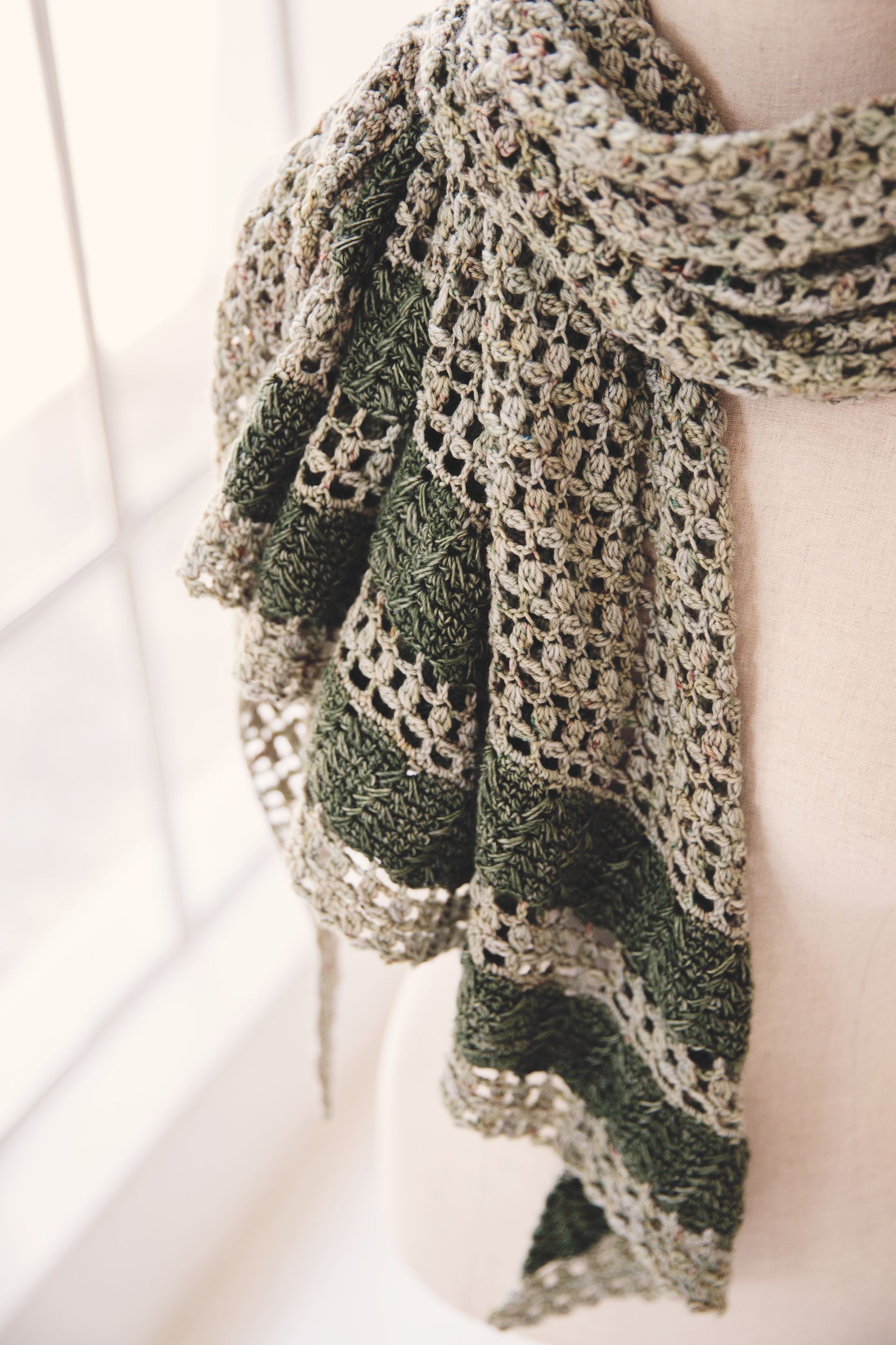 Crocheted scarf draped; shades of green and beige; openwork design with textured border.