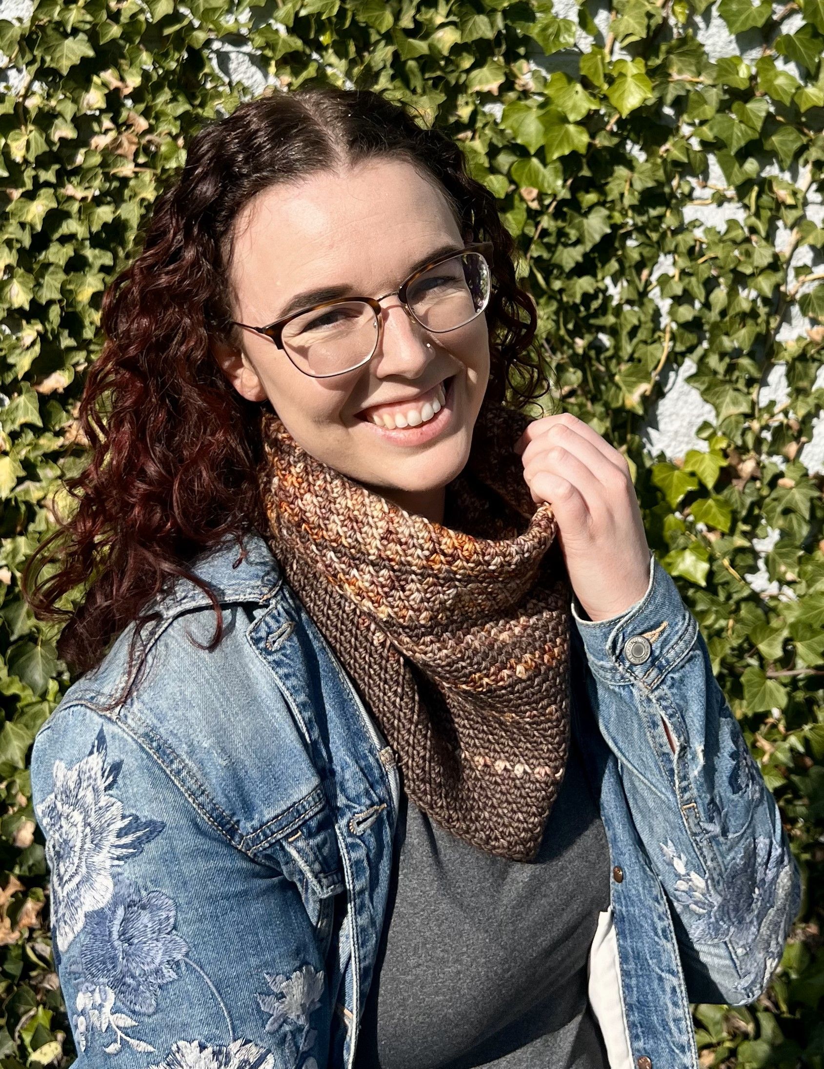 Woman with glasses smiling, wearing a denim jacket and knitted cowl, in front of ivy.